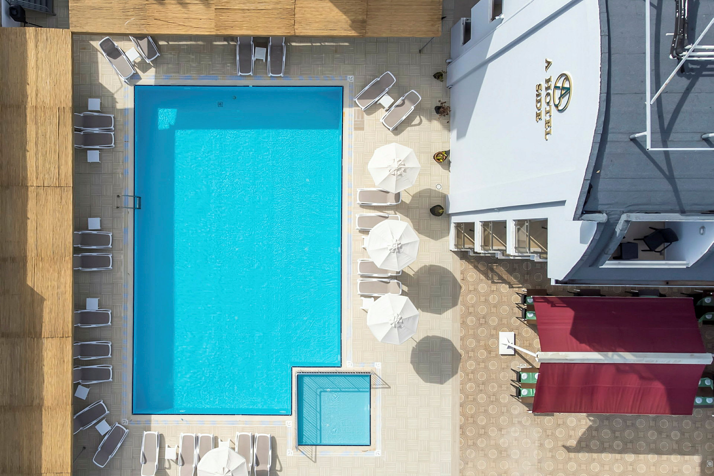Top-view af hotelpool med liggestole og parasoller, omgivet af bygninger. (AI-genereret tekst)