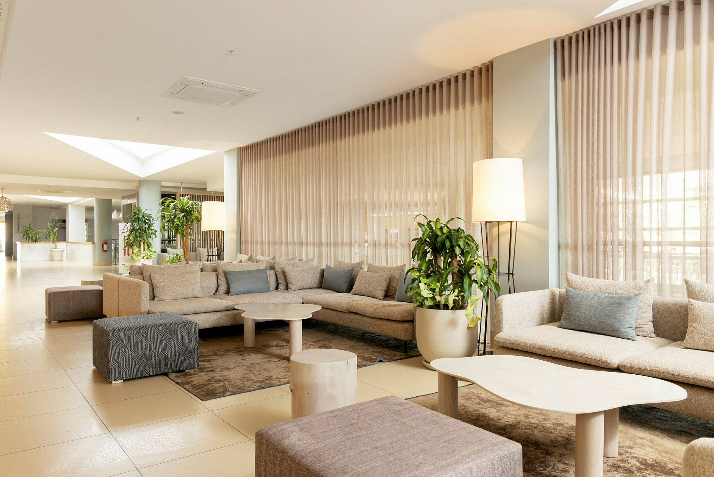 Et hotelværelse med moderne design, indeholder en gruppe komfortable beige sofaer, lave borde og grønne planter. (AI-genereret tekst)