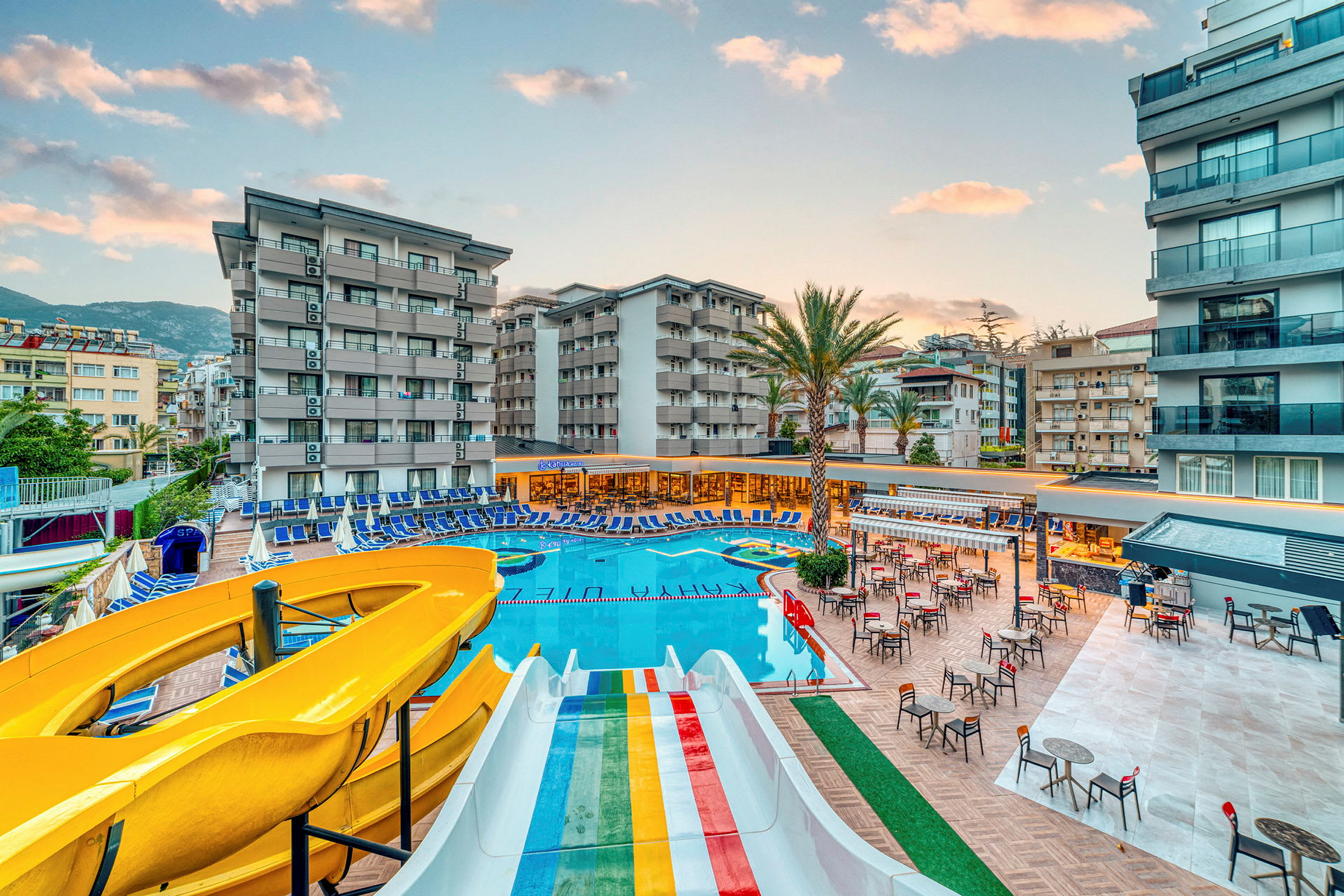 Stor gul og regnbuefarvet vandrutsjebane ved en pool omgivet af hotelbygninger og liggestole. (AI-genereret tekst)