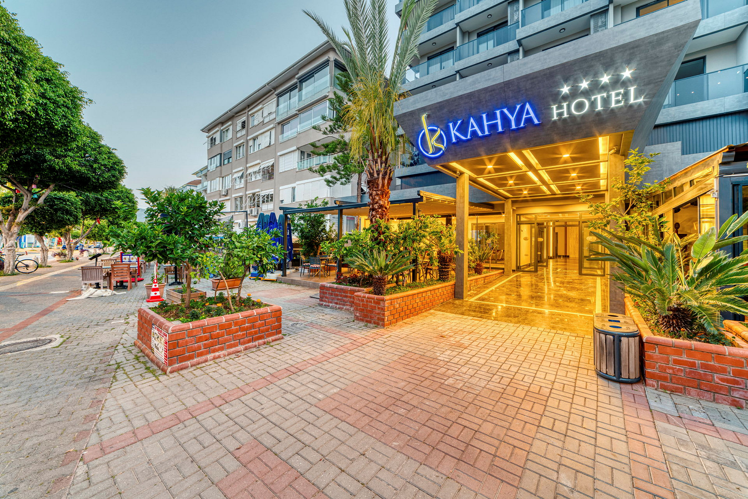 Indgangen til Kahya Hotel med omkringliggende frodige beplantninger og moderne bygninger i baggrunden. (AI-genereret tekst)