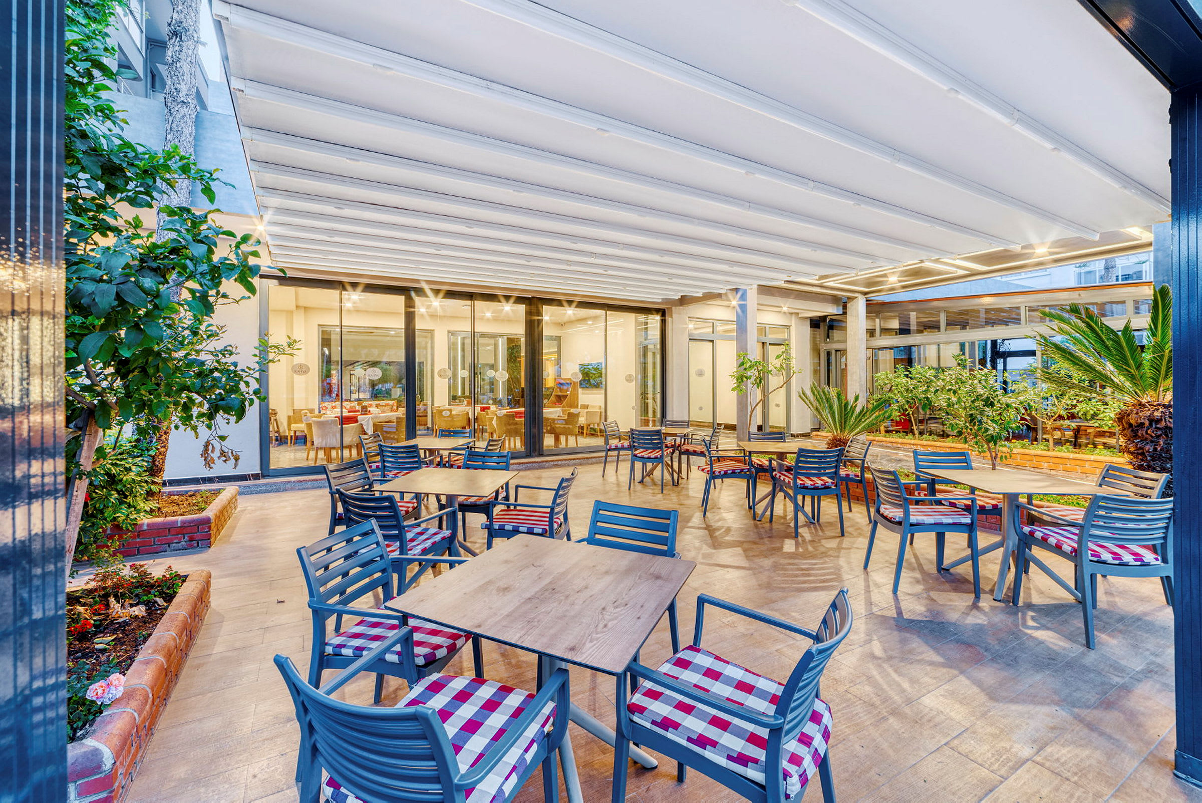 Billede af en udendørs terrasse med borde og stole samt planter og et glaseret indendørsområde i baggrunden. (AI-genereret tekst)