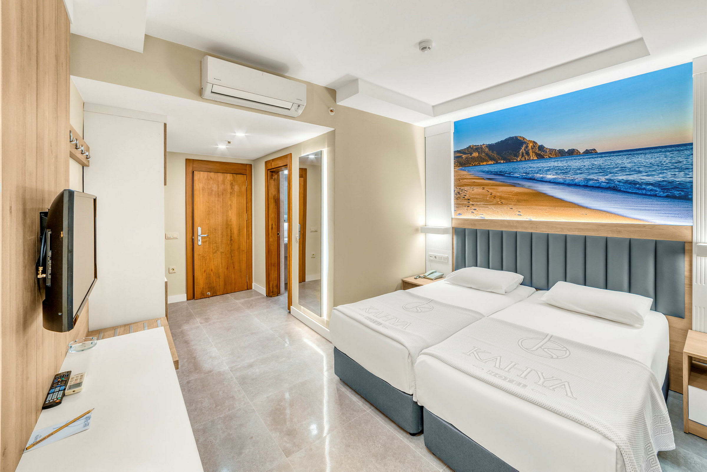 Hotelværelse med to senge, et vægmonteret TV og et dekorativt billede af en strand bag sengene. (AI-genereret tekst)
