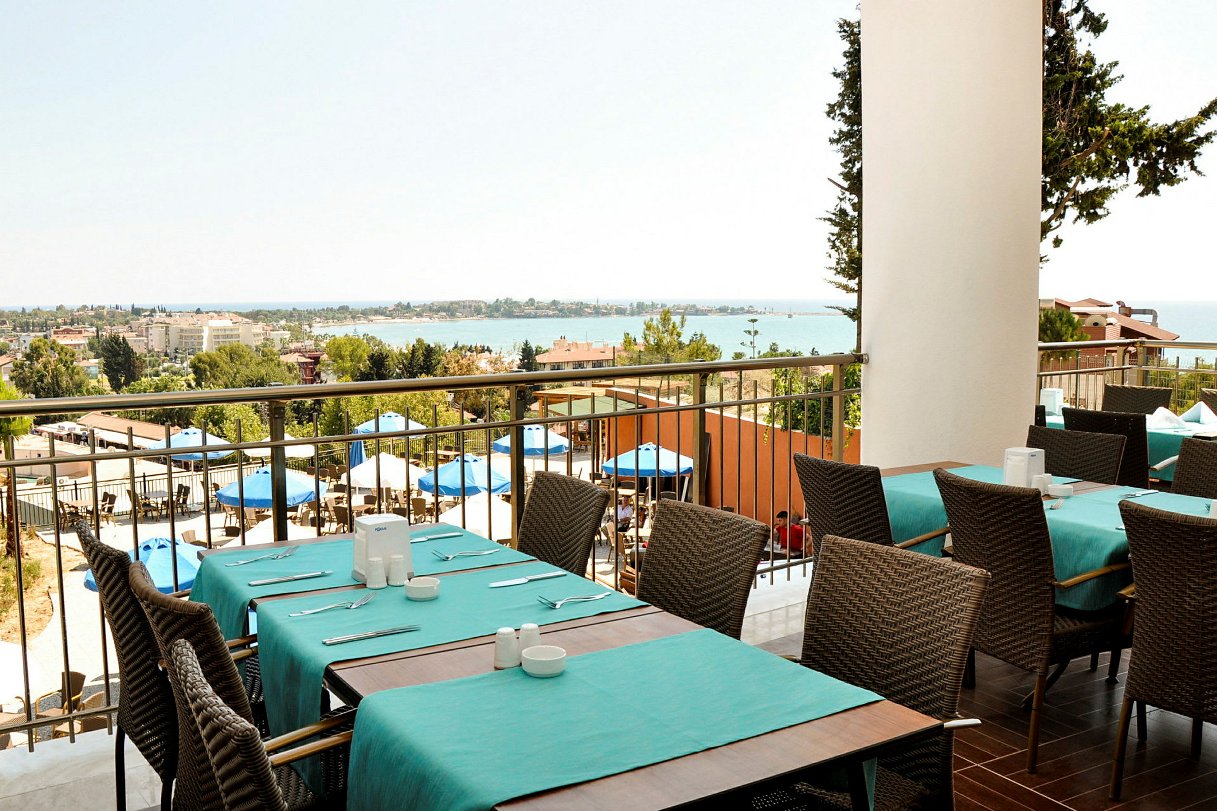 Udsigt fra en restaurant med dækkede borde og stole, udendørsområde med parasoller og havet i baggrunden. (AI-genereret tekst)
