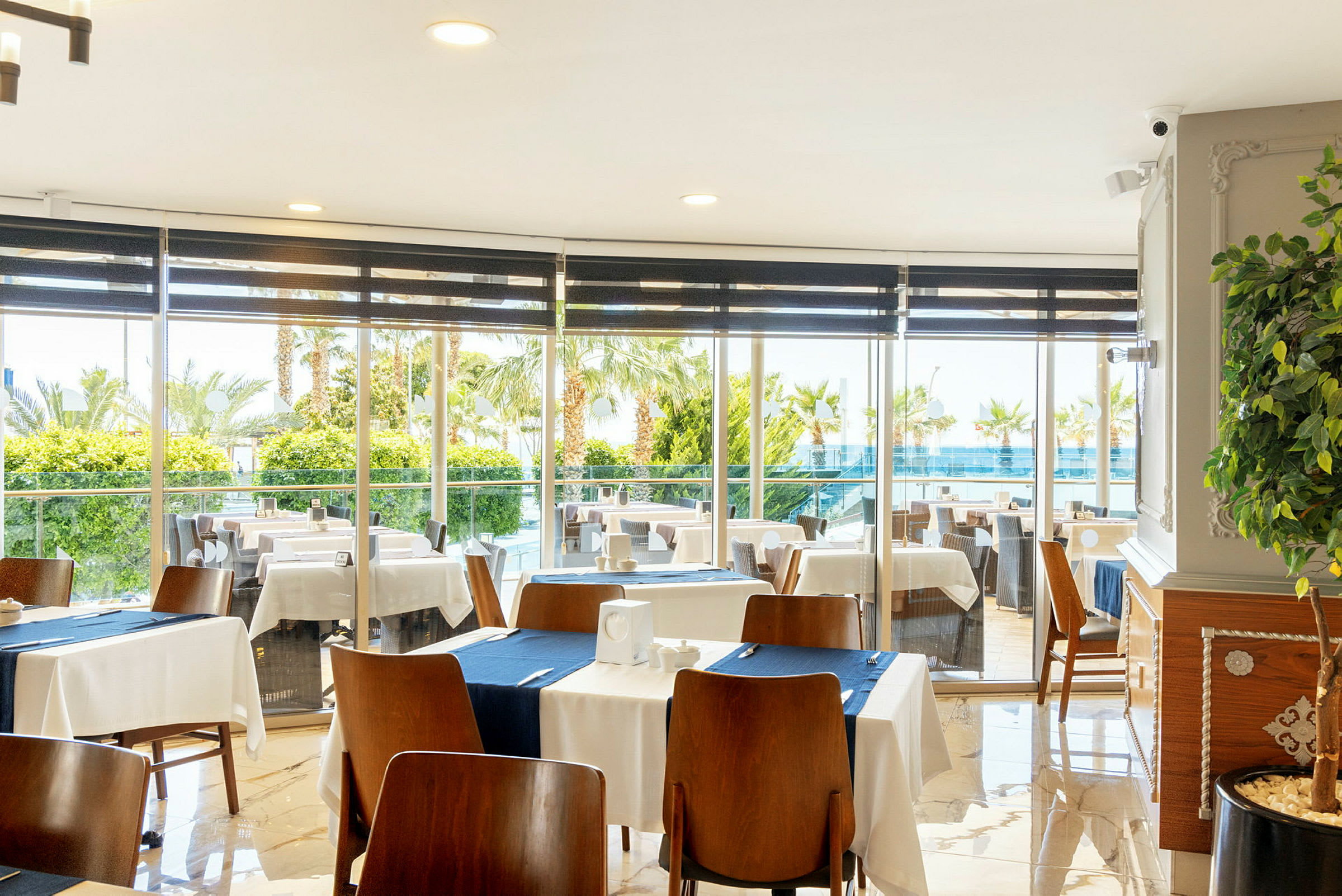 Restaurant med dækkede borde og udsigt gennem vinduer mod grønt område og pool. (AI-genereret tekst)