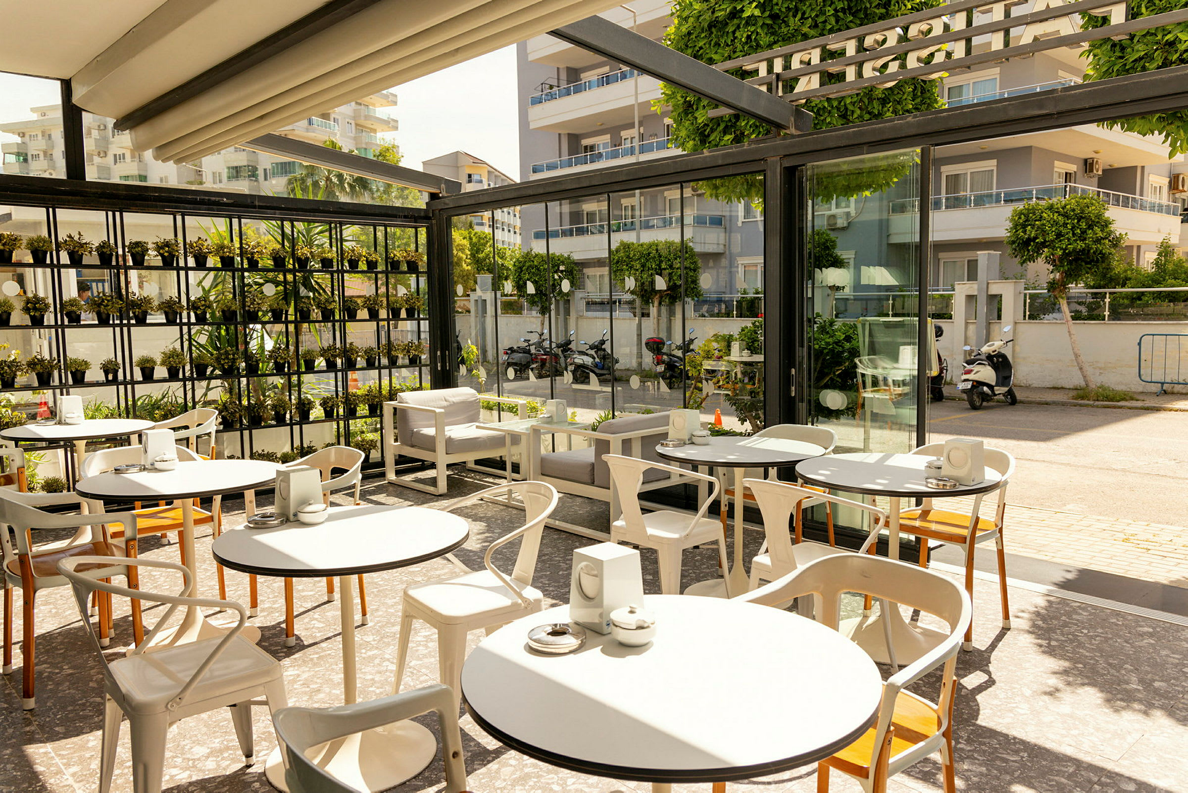 En pæn caféterrasse med borde og stole, omgivet af glasvægge og grønne planter. (AI-genereret tekst)