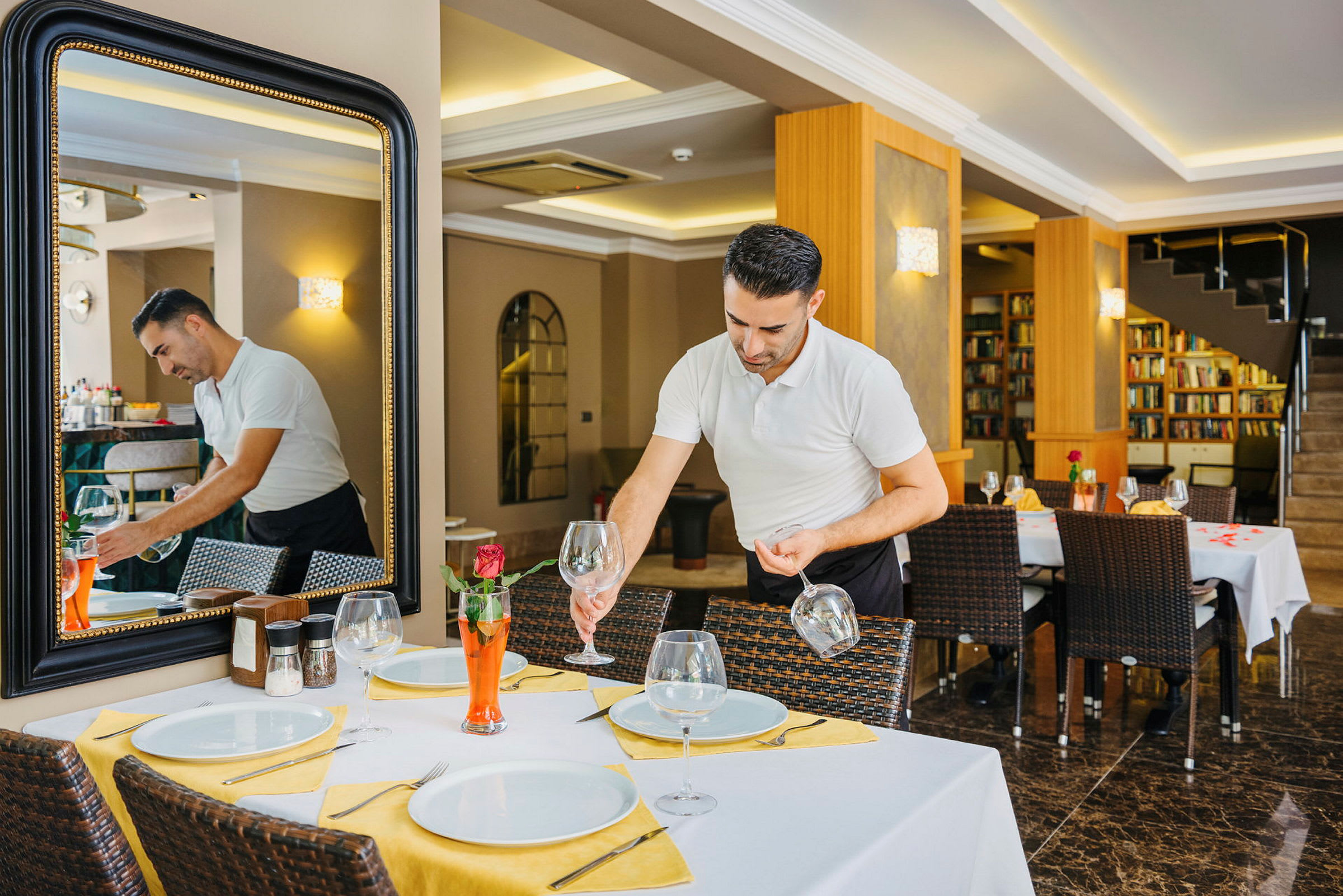 En person dækker et bord på en restaurant med hvide tallerkener, glas og gul borddekoration. (AI-genereret tekst)