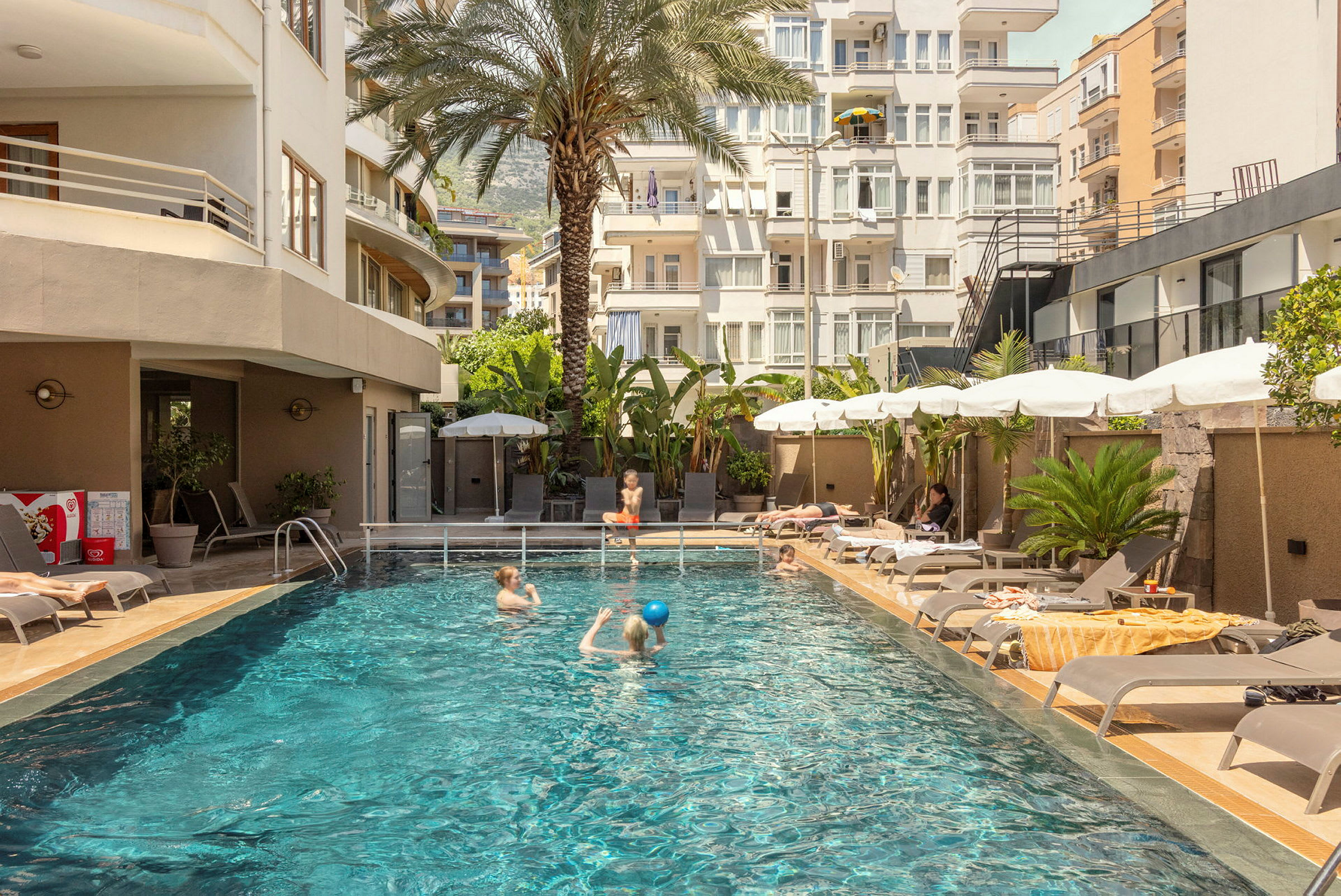 Billede af en udendørs pool omgivet af liggestole og parasoller samt omkringliggende bygninger. (AI-genereret tekst)