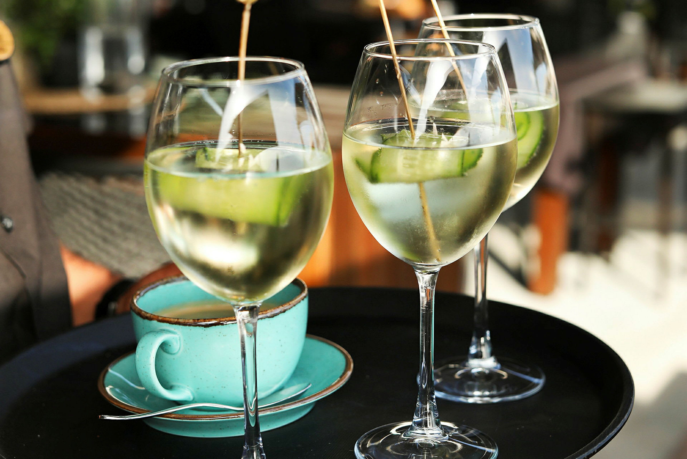 Tre glas med drinks indeholdende isterninger og agurkeskiver samt en turkis kaffekop på en serveringsbakke. (AI-genereret tekst)