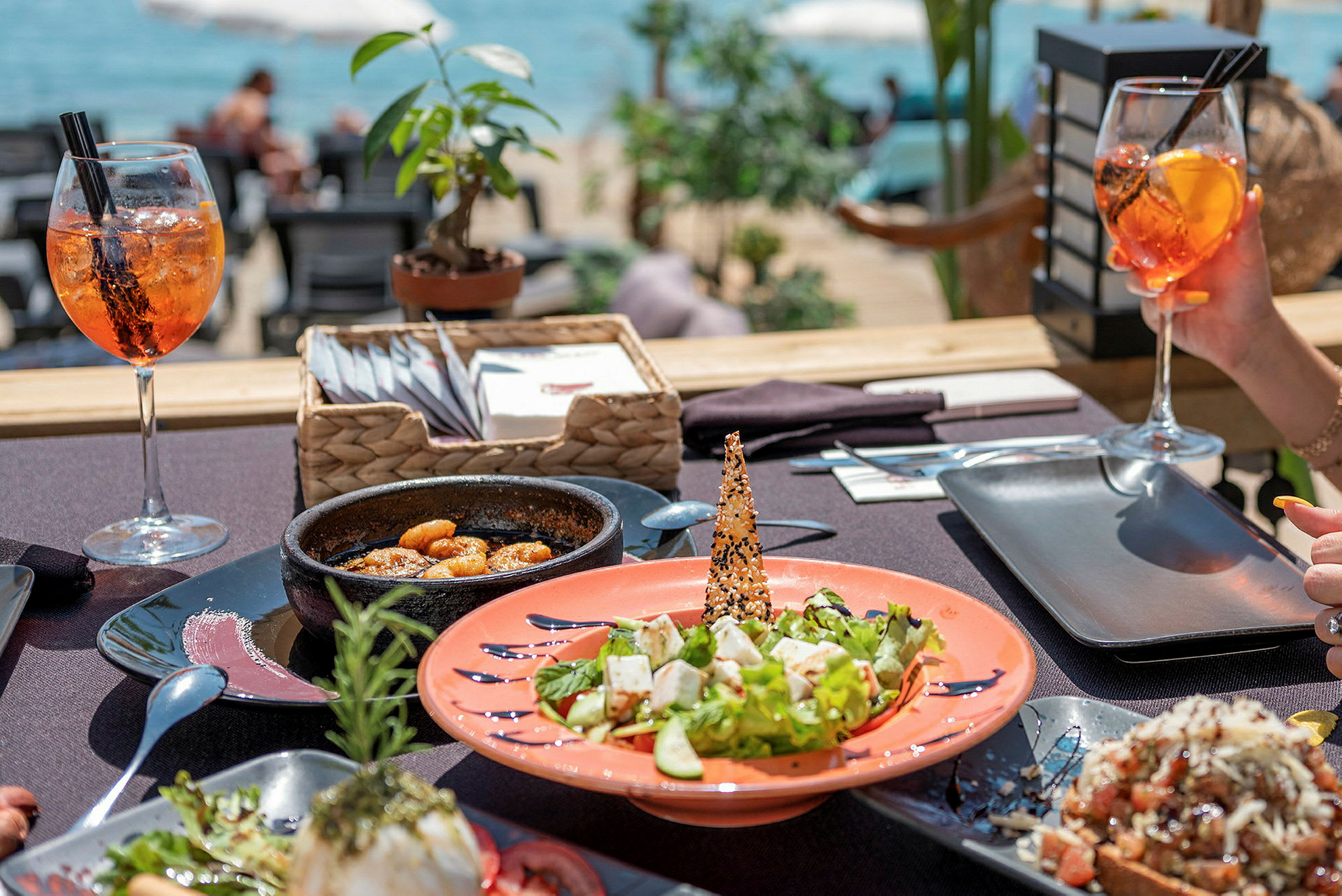 Et bord med forskellige retter og drikkevarer, der er opstillet på en udendørsrestaurant ved havet. (AI-genereret tekst)