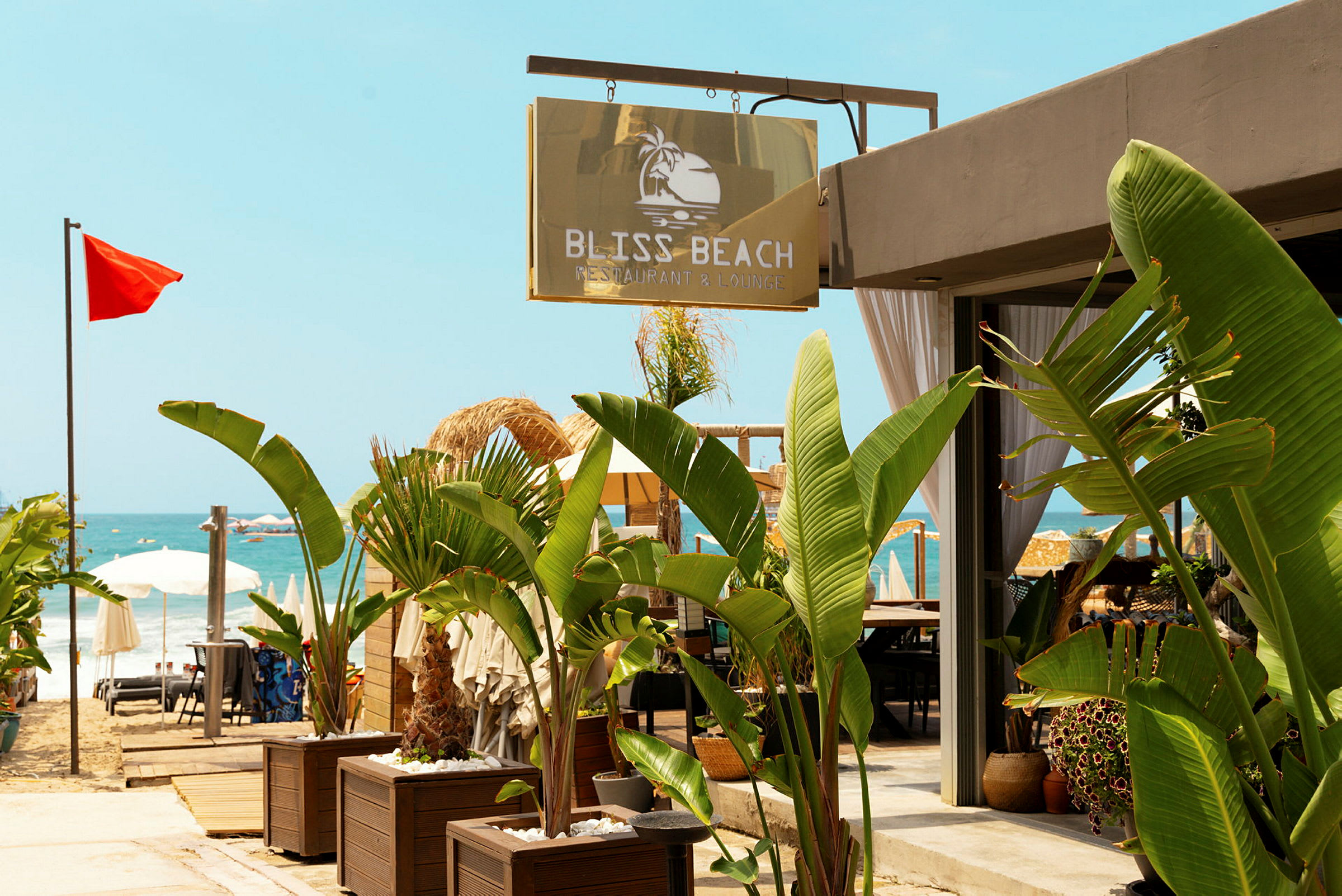 Skilt med teksten Bliss Beach Restaurant & Lounge ved en strand med røde flag og parasoller. (AI-genereret tekst)