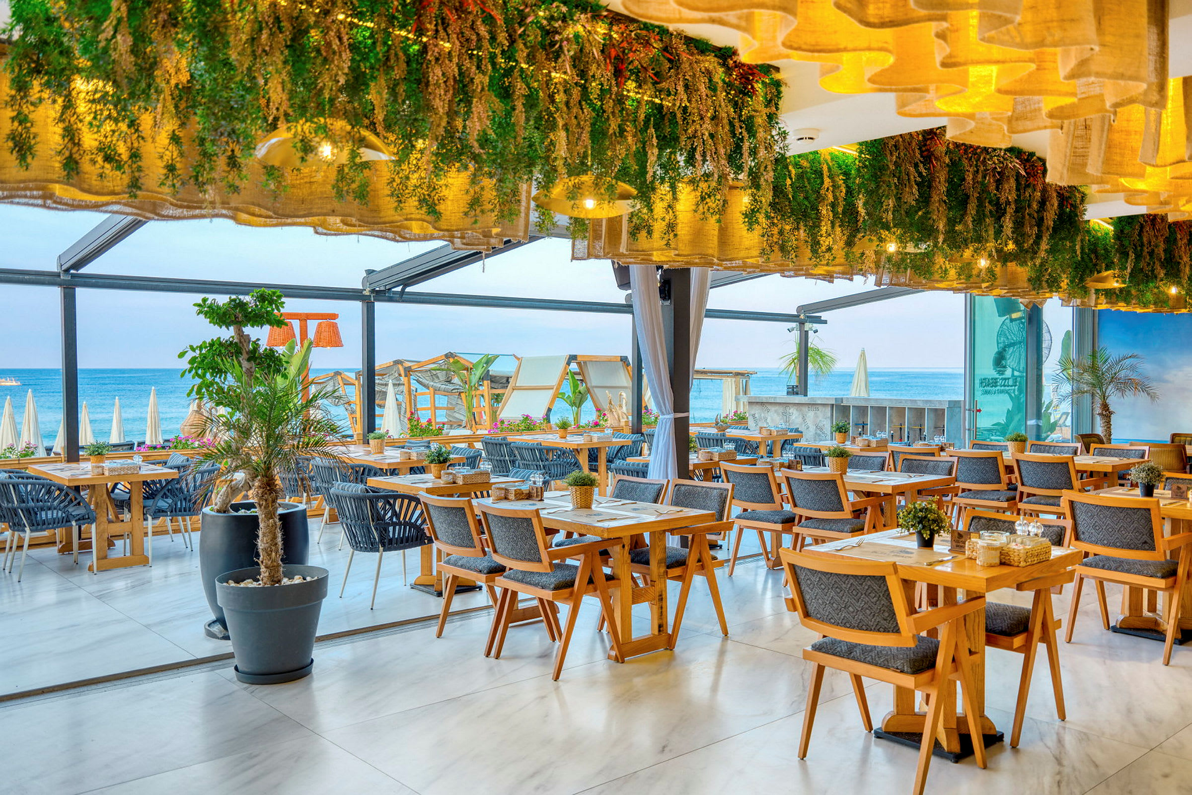 Restaurant med træborde, dekorative planter og udsigt til stranden. (AI-genereret tekst)