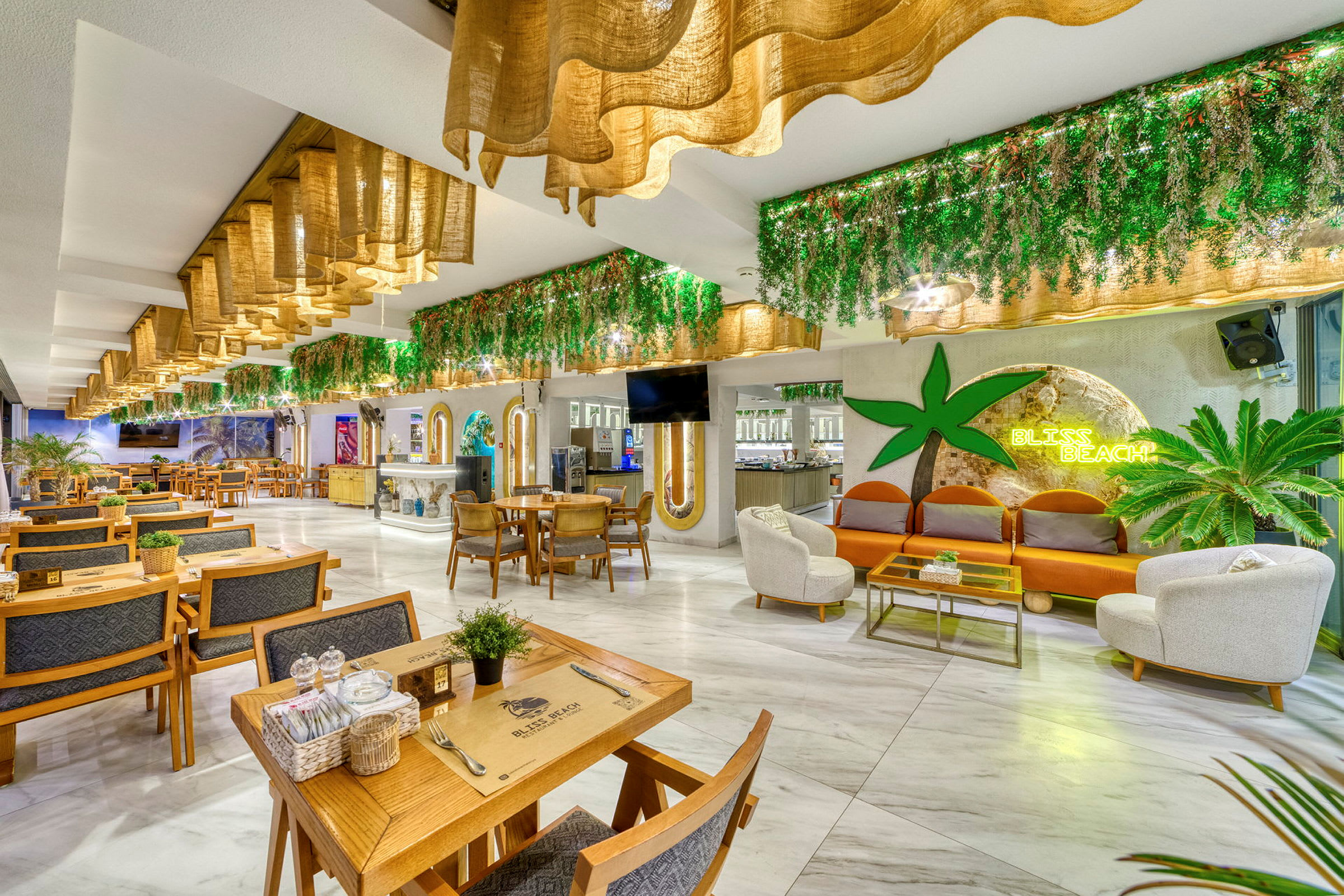 Interiør af en restaurant med siddegrupper, grønne dekorationer og et skilt, der lyser med teksten Bliss Beach. (AI-genereret tekst)