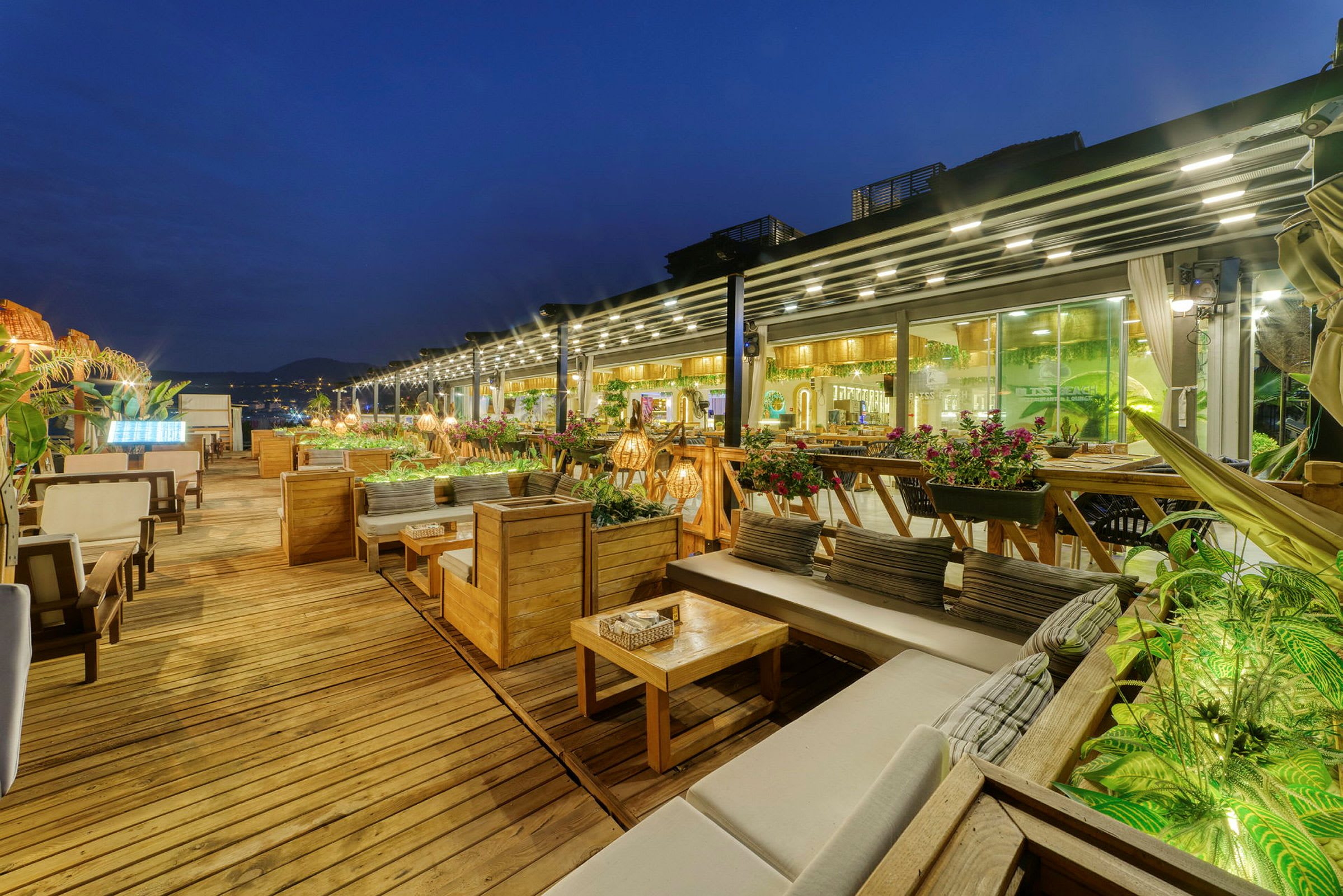 En udendørsrestaurant med træterrasse, sofaer og borde, samt grønne planter og lyskæder. (AI-genereret tekst)