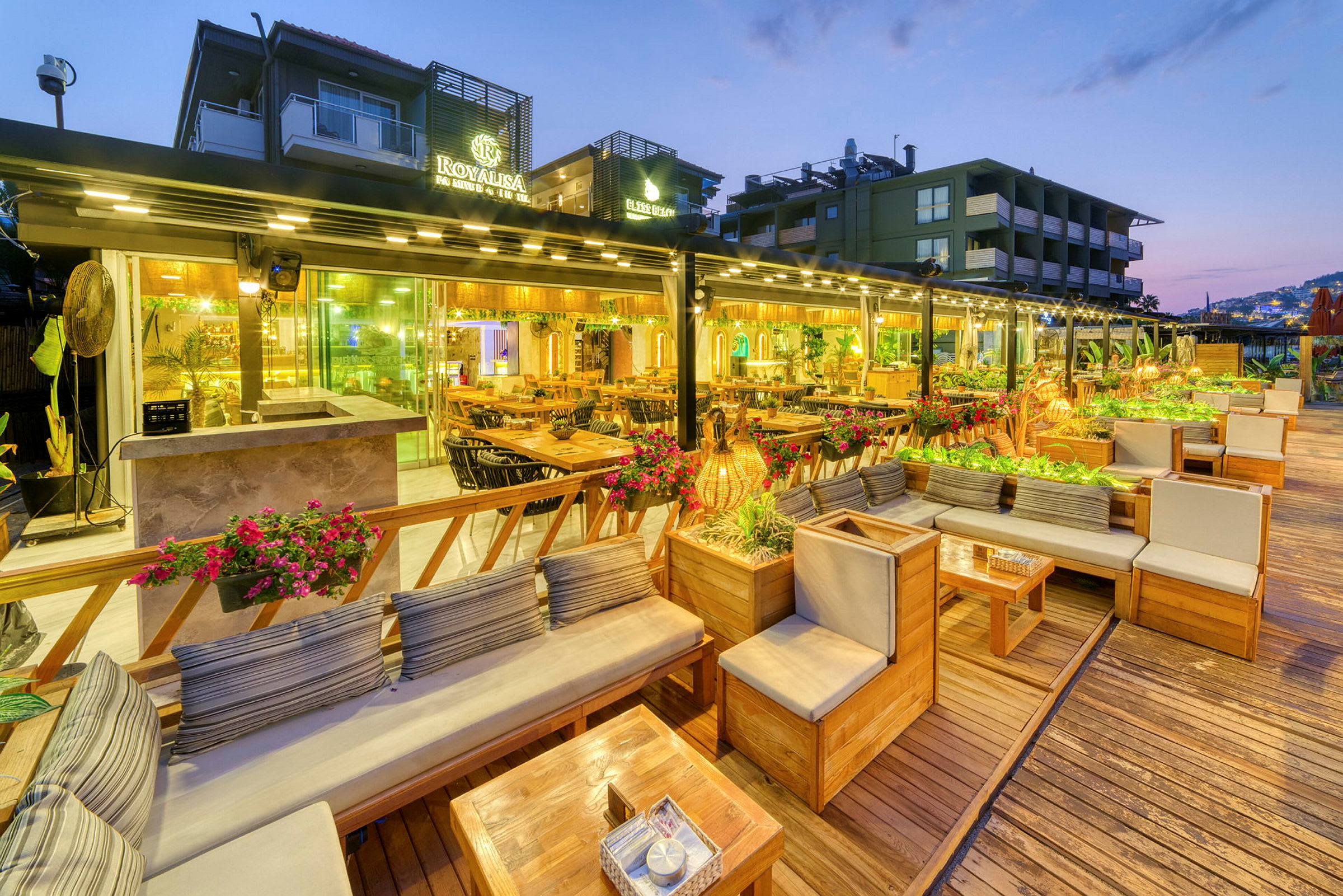 Billede af en udendørs terrasse med siddepladser og borde, omgivet af planter og dekorationer, belyst om aftenen. (AI-genereret tekst)