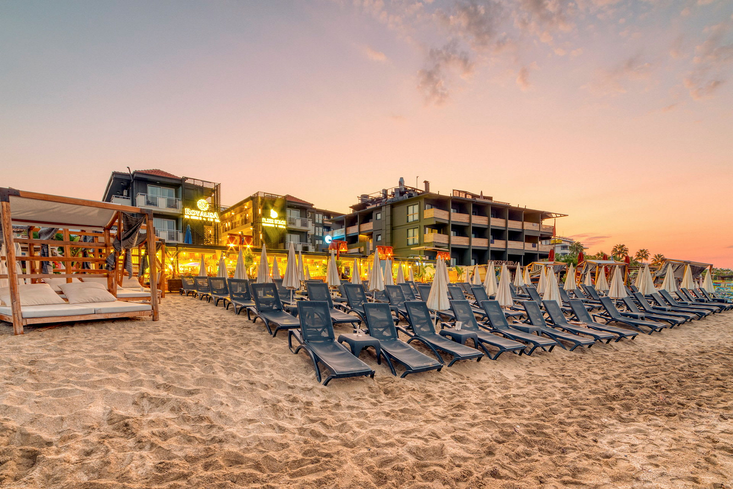 Solstole og parasoller på en sandstrand med hotel i baggrunden ved solnedgang. (AI-genereret tekst)