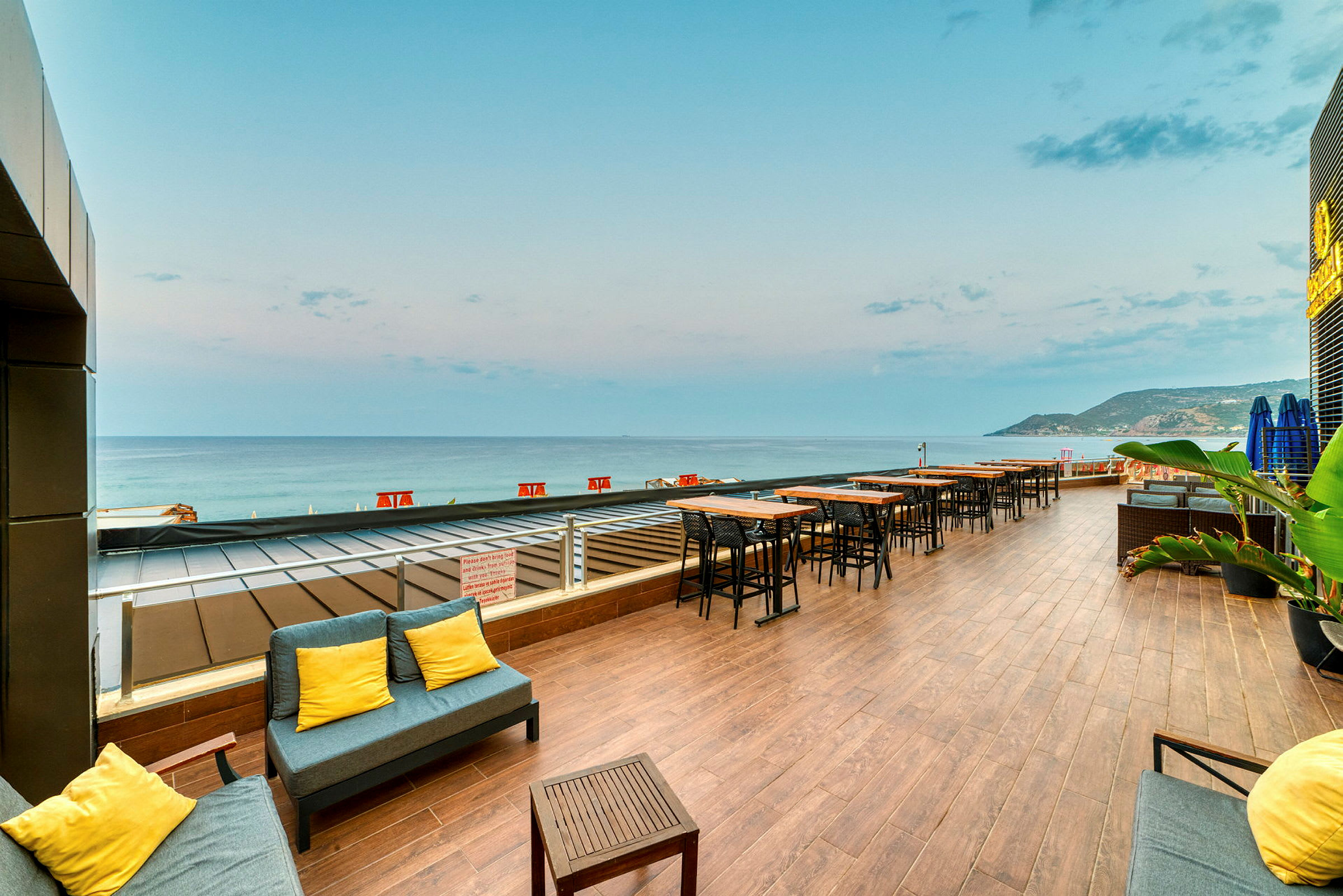 Udendørs terrasse med borde og stole samt sofa med gule puder, med udsigt over havet. (AI-genereret tekst)