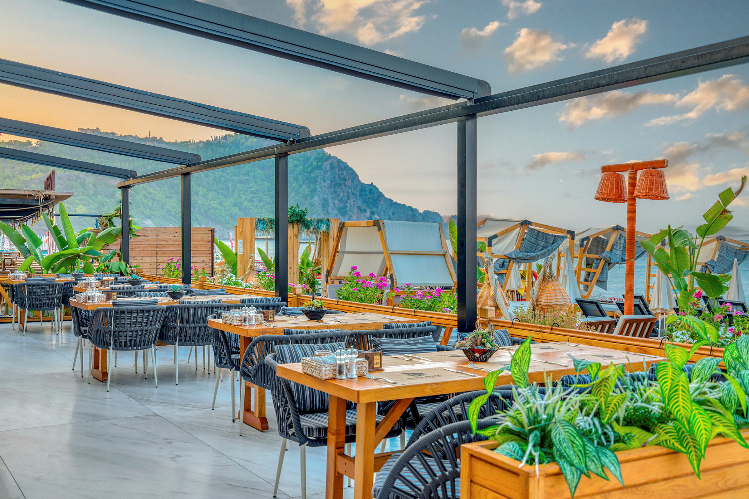 Udendørsrestaurant med borde og stole, omgivet af planter og med udsigt over havet og bjergene. (AI-genereret tekst)