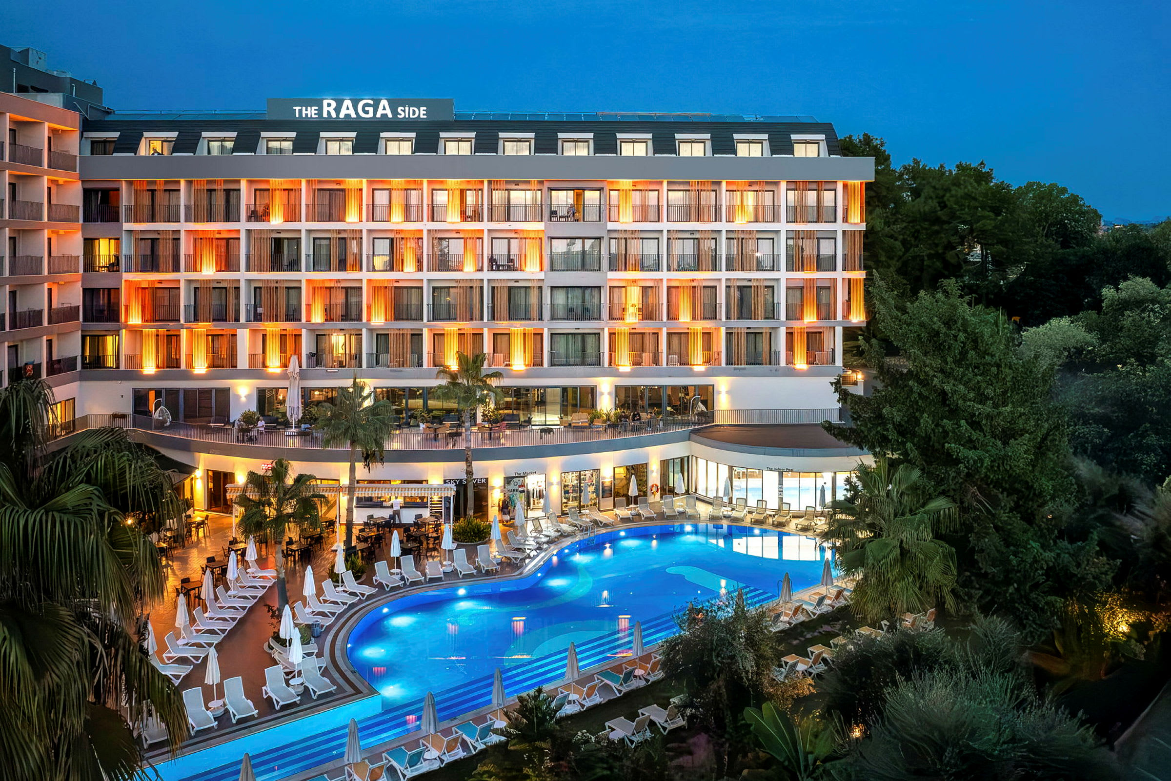 Hotellets facade i skumringen med oplyste altaner og en pool omgivet af liggestole. (AI-genereret tekst)