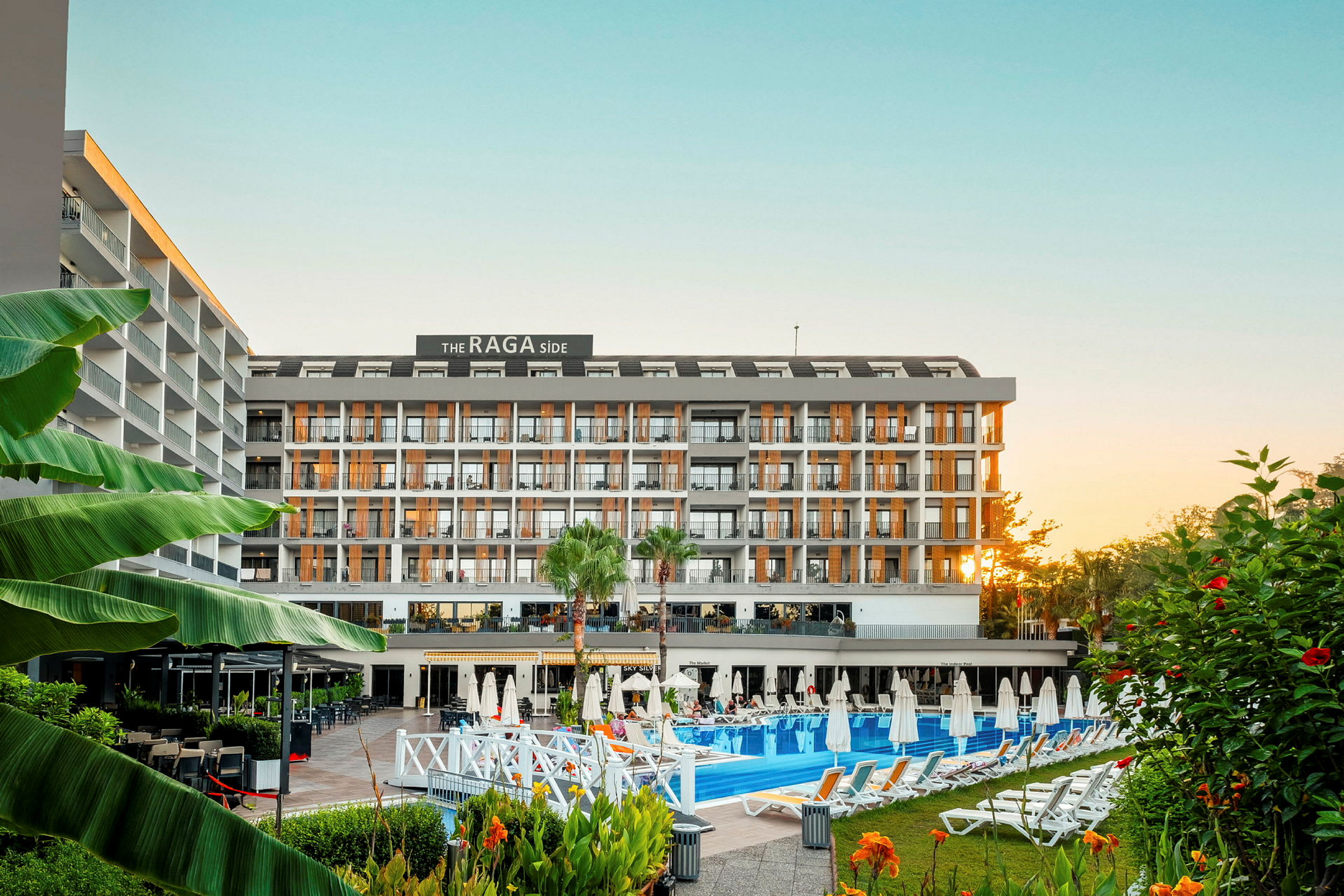 Et fler-etagers hotelkompleks set forfra, omgivet af swimmingpool, solsenge og beplantning. (AI-genereret tekst)
