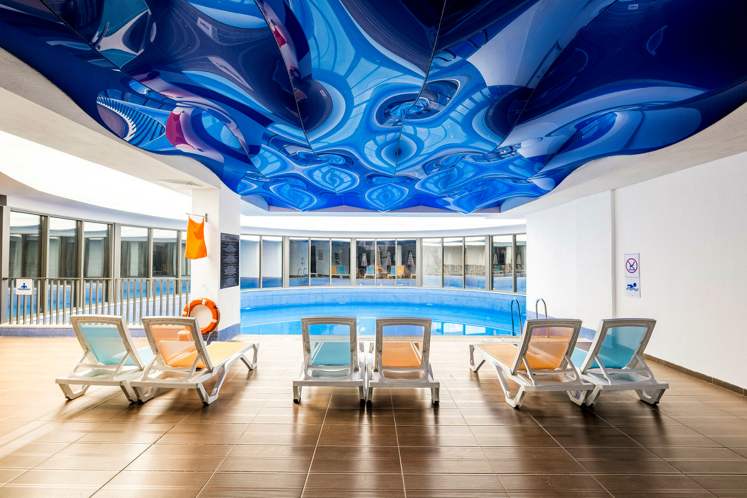 Indendørs swimmingpool med flere liggestole og et blåt unikt mønstret loft. (AI-genereret tekst)