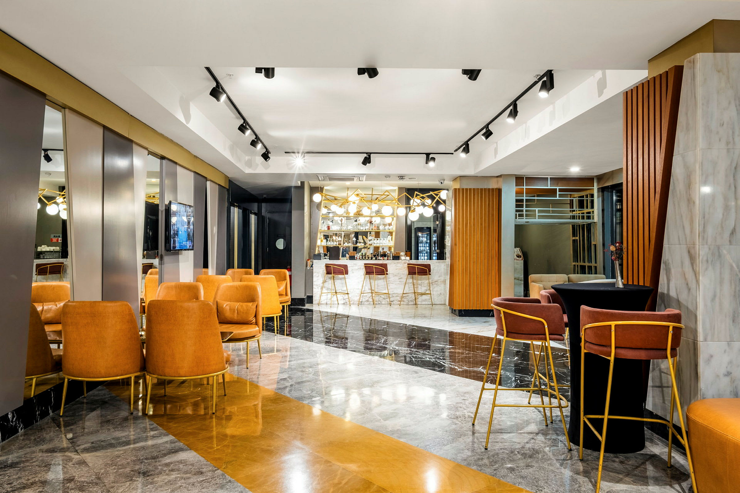 Interiøret viser en moderne lounge med siddepladser, sofaer og en bar i baggrunden udsmykket med lysekroner og spejle. (AI-genereret tekst)