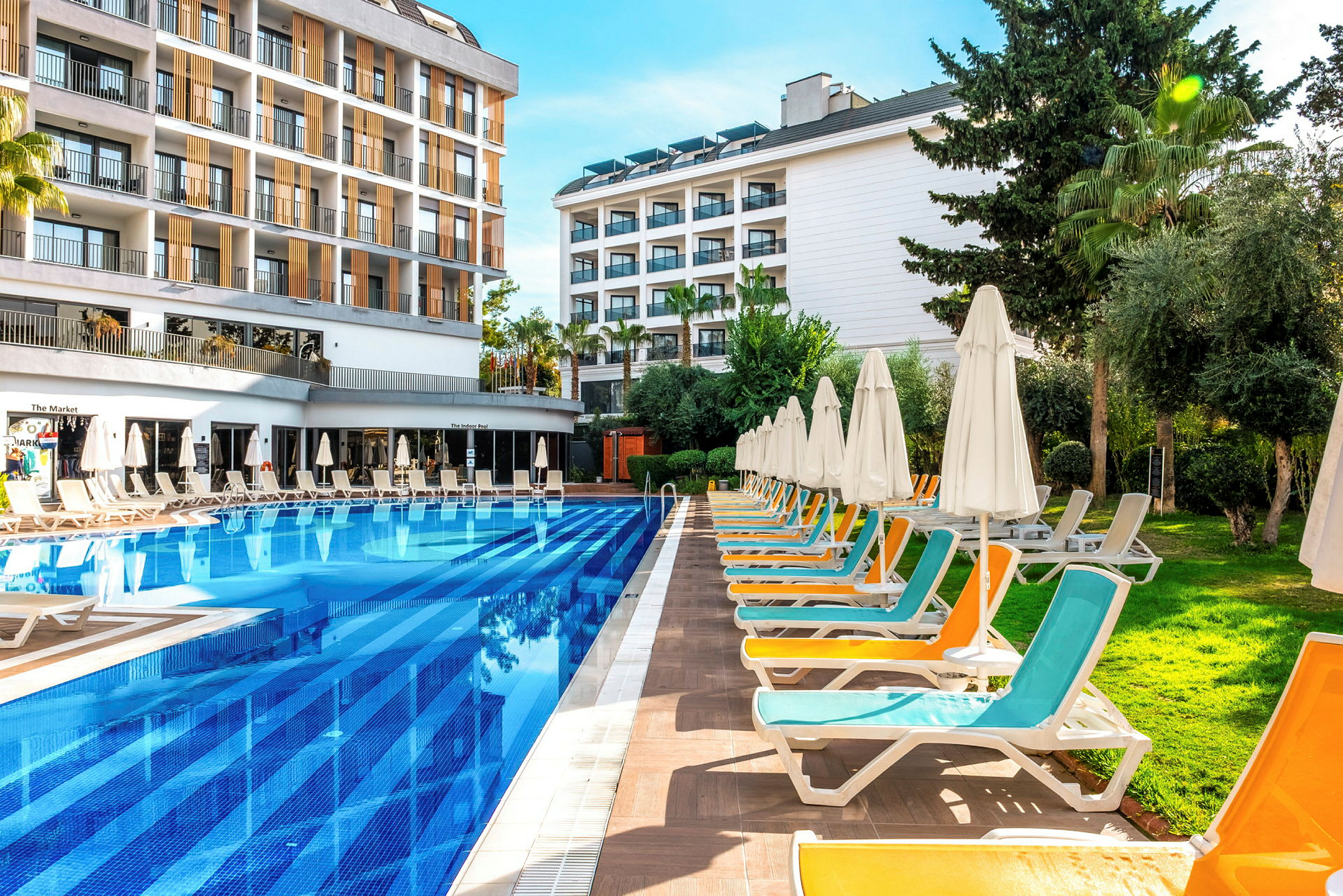 Et hotels udendørsområde med en pool, omkringliggende liggestole og parasoller samt bygninger i baggrunden. (AI-genereret tekst)