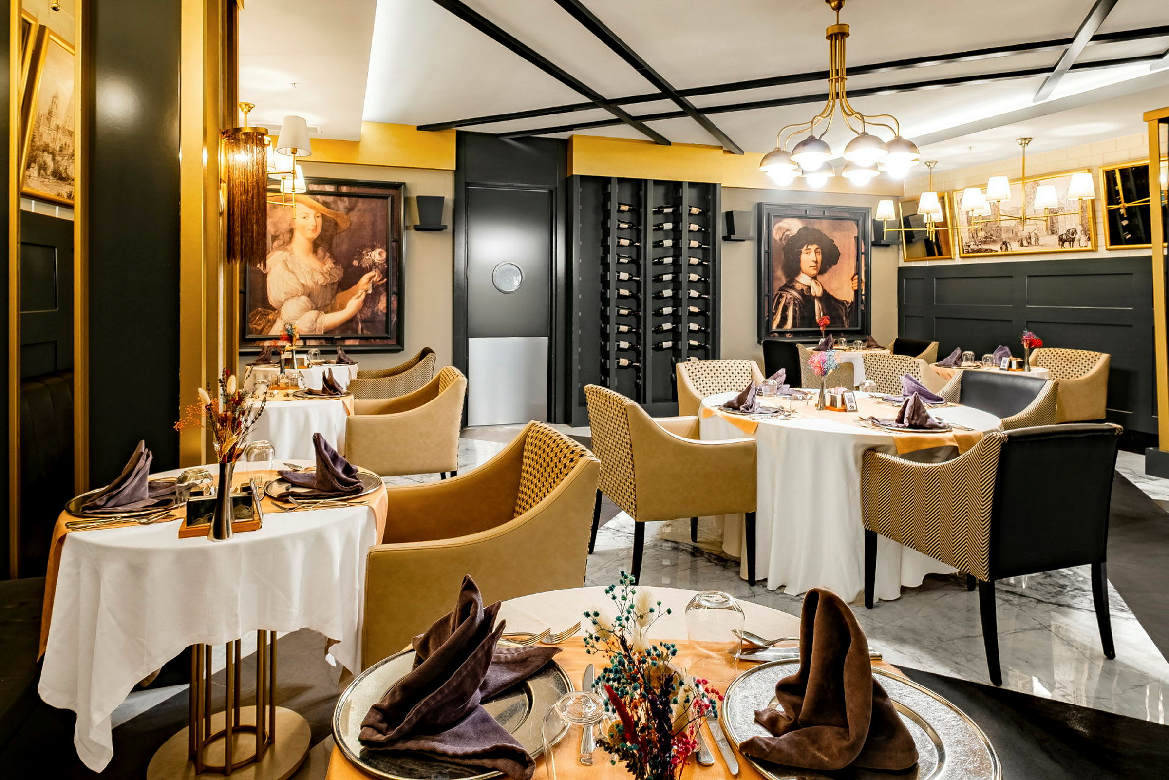 Et elegant indrettet restaurantområde med runde borde dækket med hvide duge, omgivet af moderne stole og dekorationer, herunder kunstværker på væggene og en vinhylde. (AI-genereret tekst)