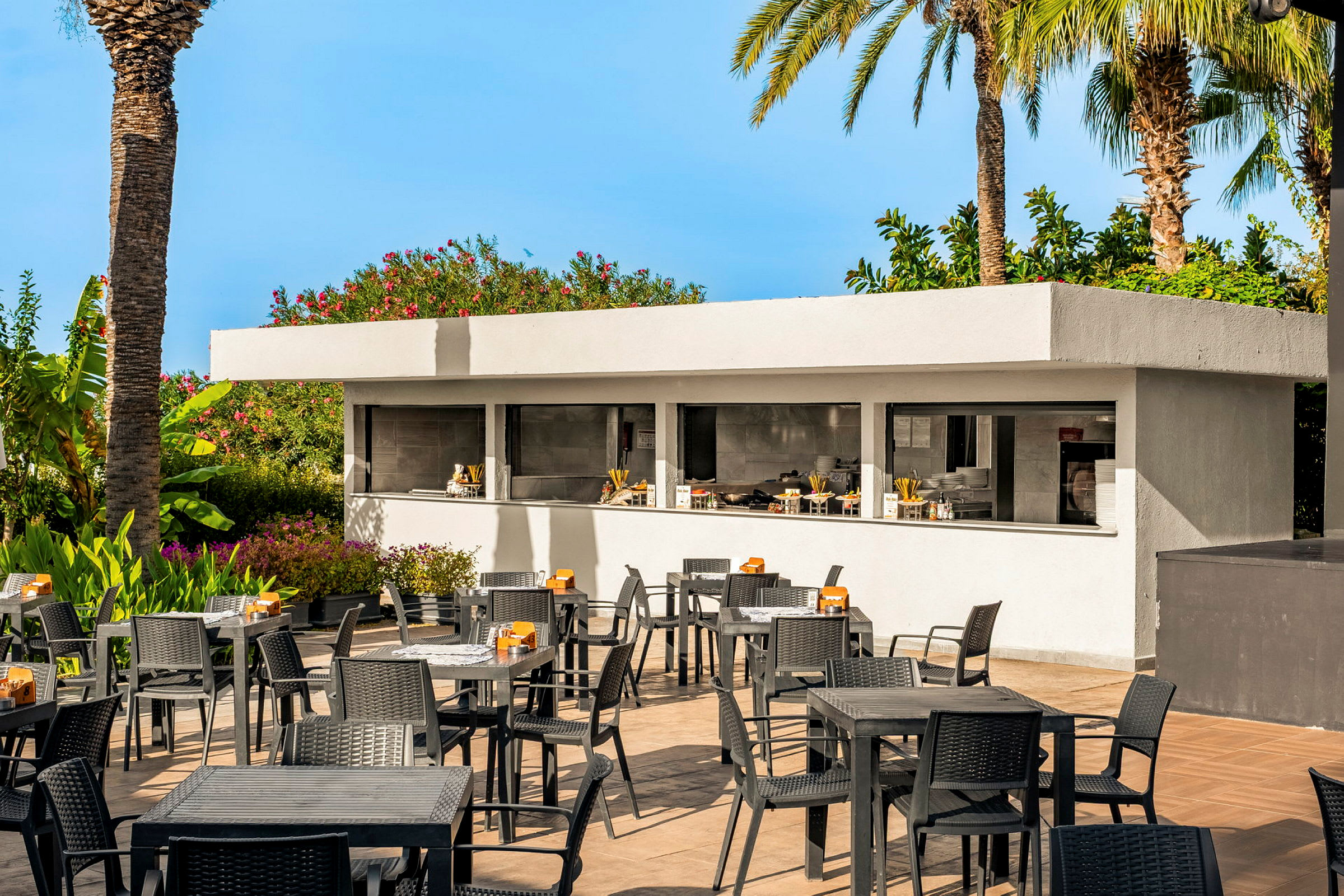 Et udendørsområde med borde og stole i forgrunden og en moderne serveringsbar i baggrunden omgivet af frodig vegetation og palmer. (AI-genereret tekst)