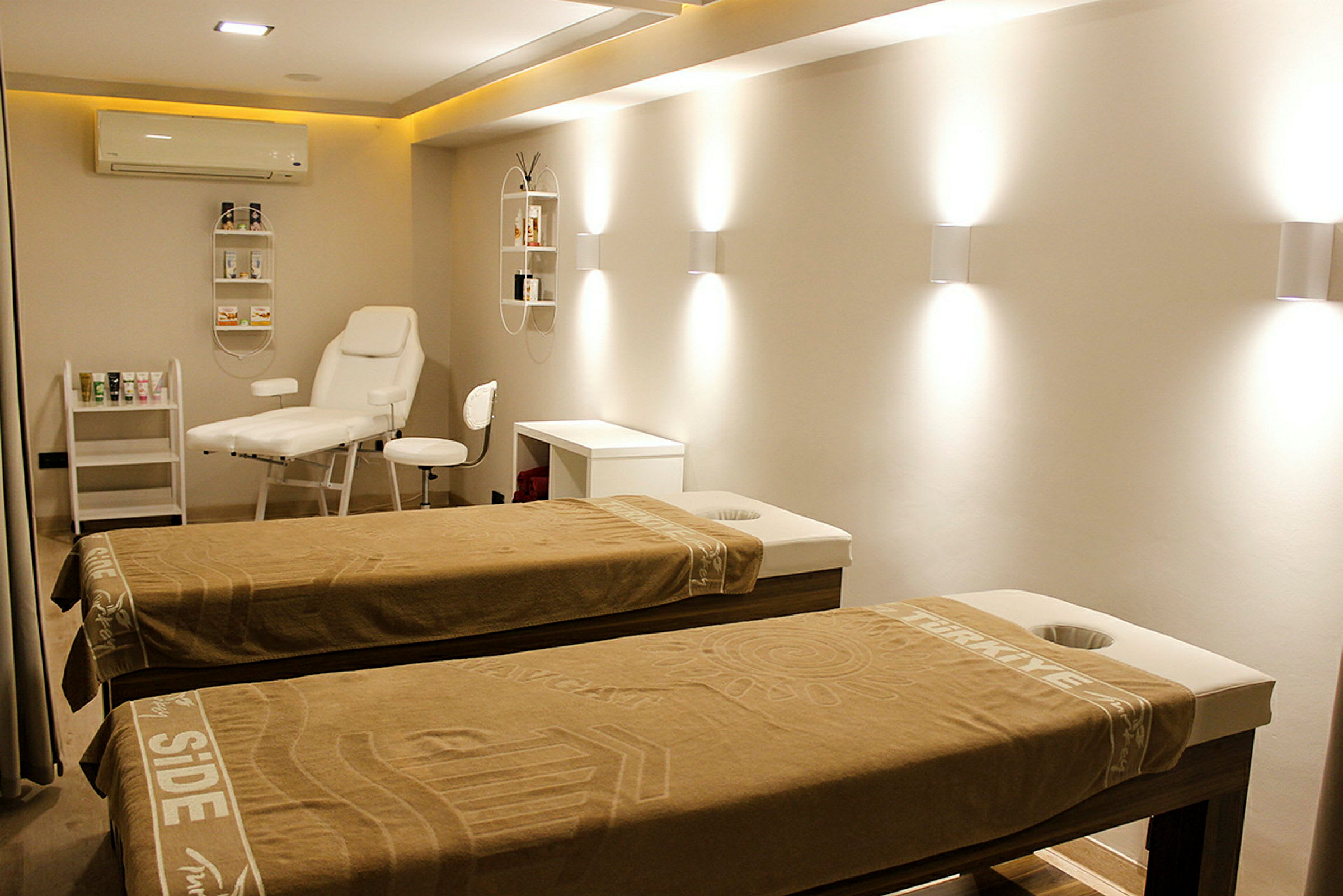 Rummet viser et lyst og moderne behandlingsområde med to massageborde dækket med brune håndklæder. På væggen hænger hylder med produkter, og der findes en hvid behandlingsstol og bord. (AI-genereret tekst)