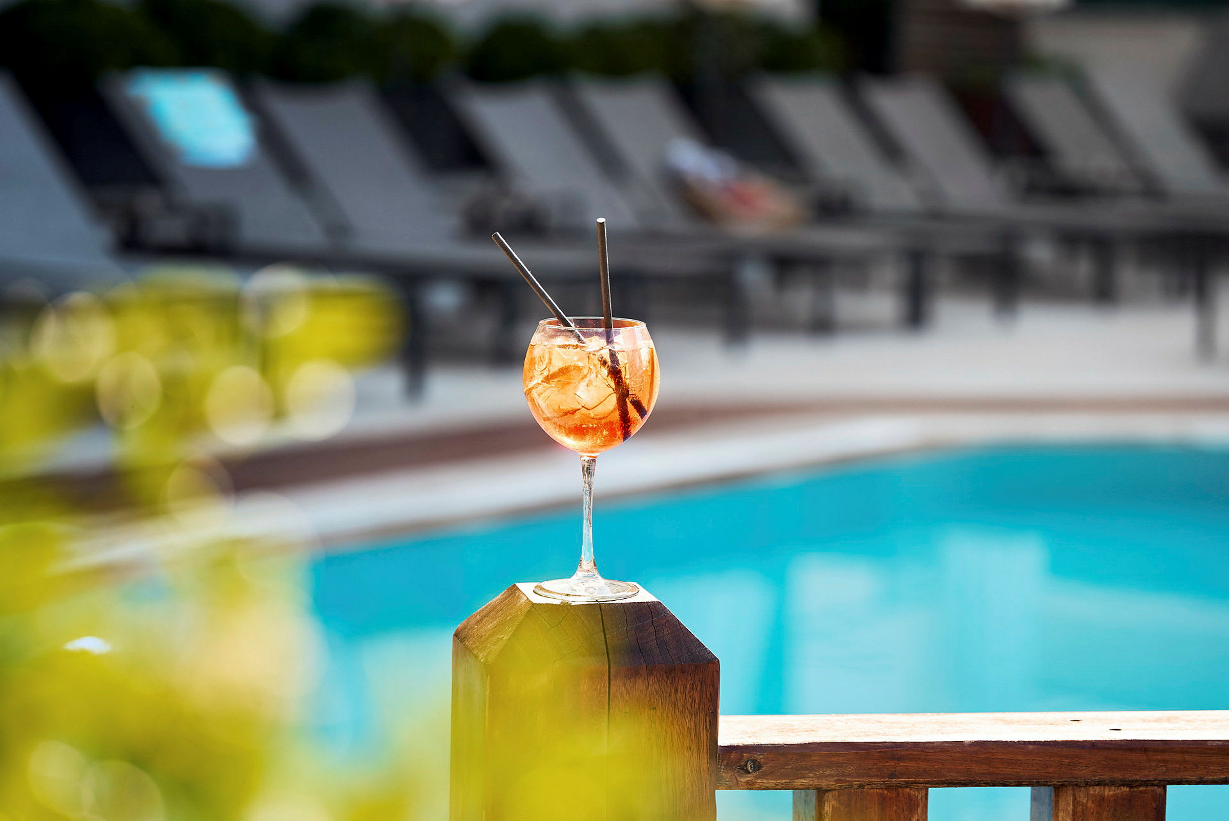 Et glas drink står på et træhegn ved en pool. (AI-genereret tekst)