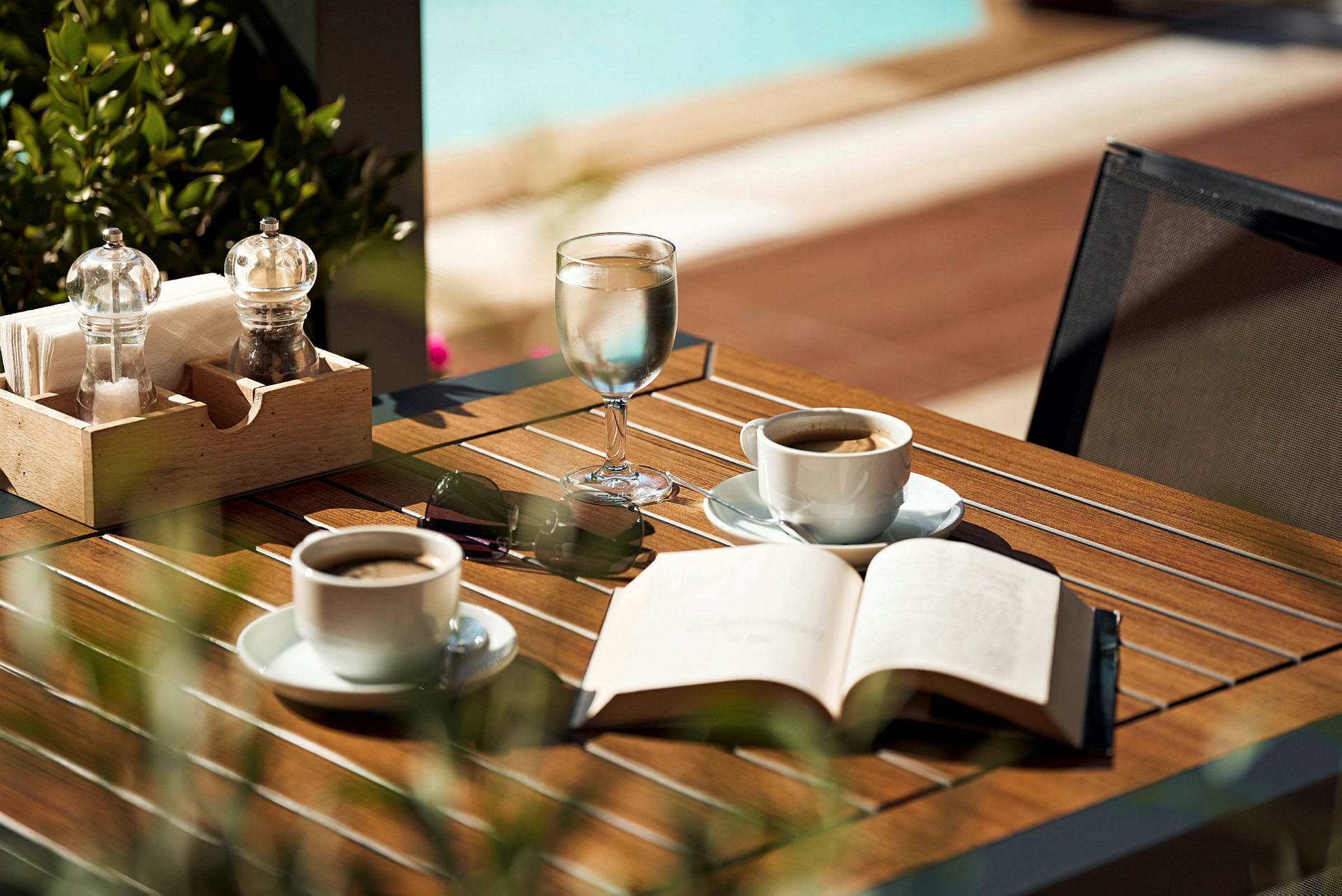Et bord med kaffekopper, et glas vand, en åben bog og solbriller. (AI-genereret tekst)