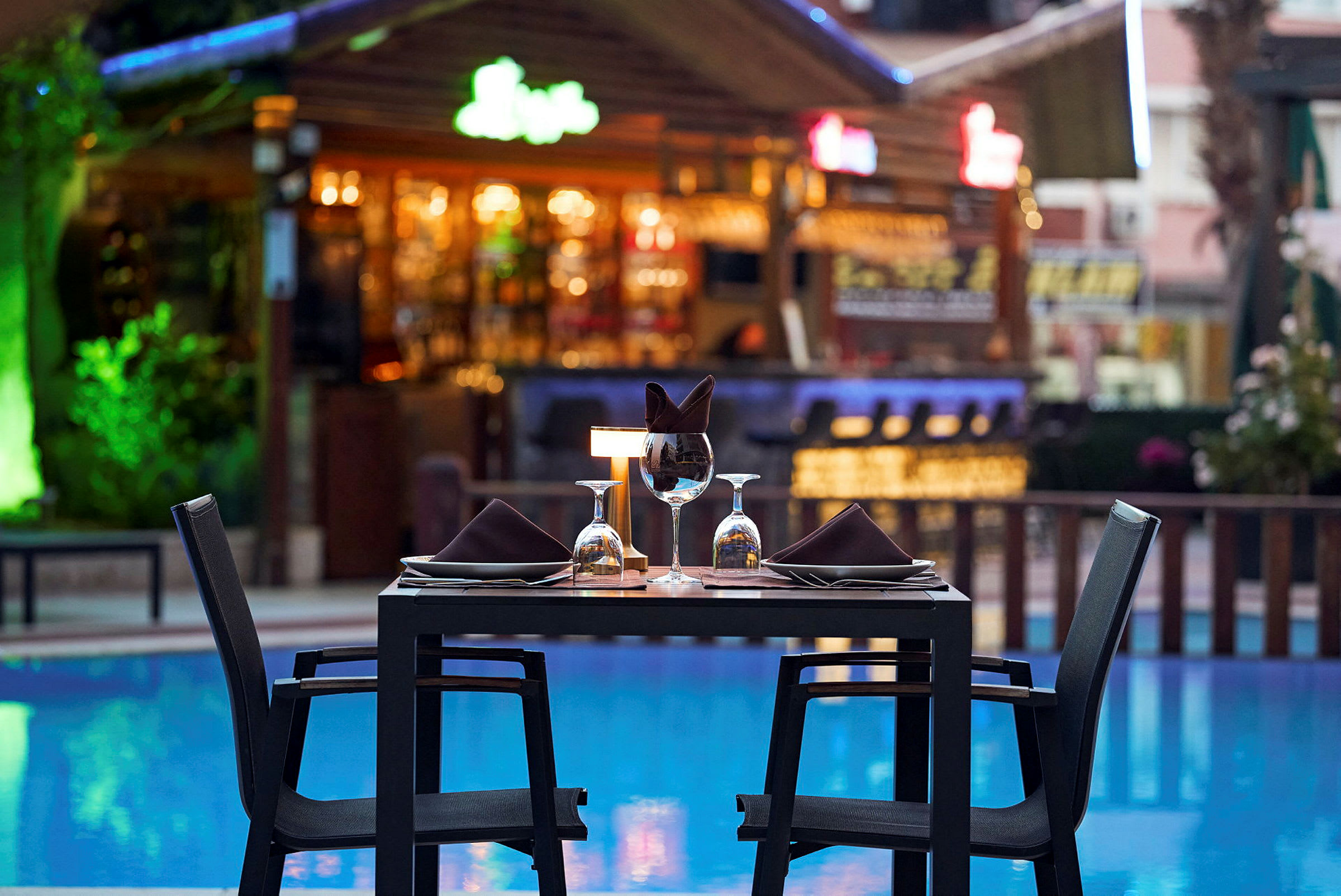 Et bord med opdækning ved en pool, i baggrunden ses en bar. (AI-genereret tekst)