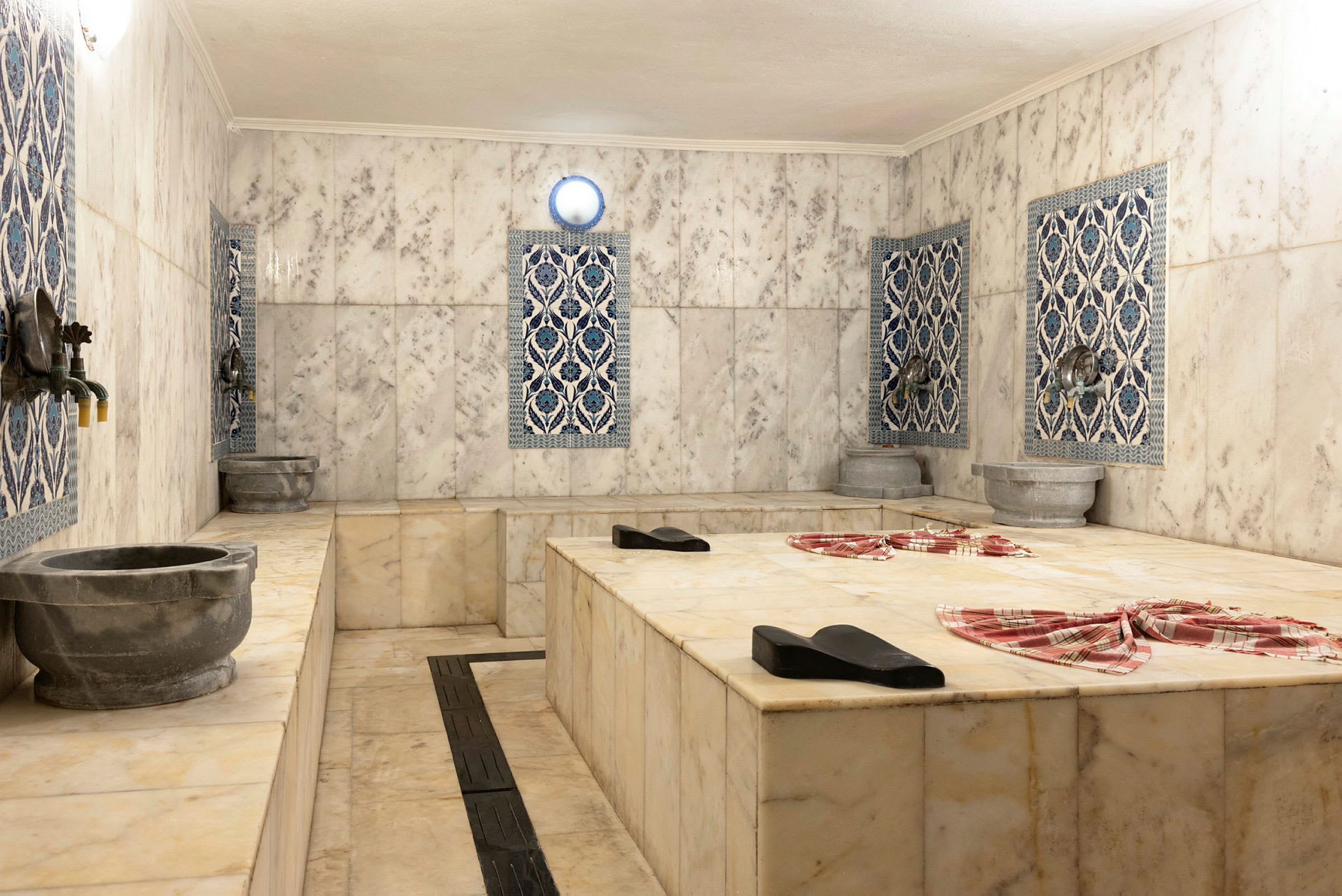 Et traditionelt hamam med marmorgulve og dekorative fliser på væggene. Stenkummer og vandhaner er synlige rundt om. (AI-genereret tekst)