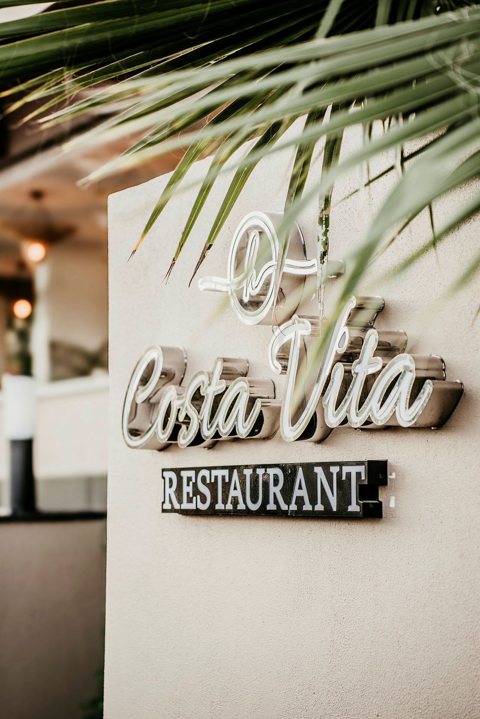Et skilt med teksten 'Costa Vita Restaurant' på en væg, omgivet af palmeblade dekoration. (AI-genereret tekst)