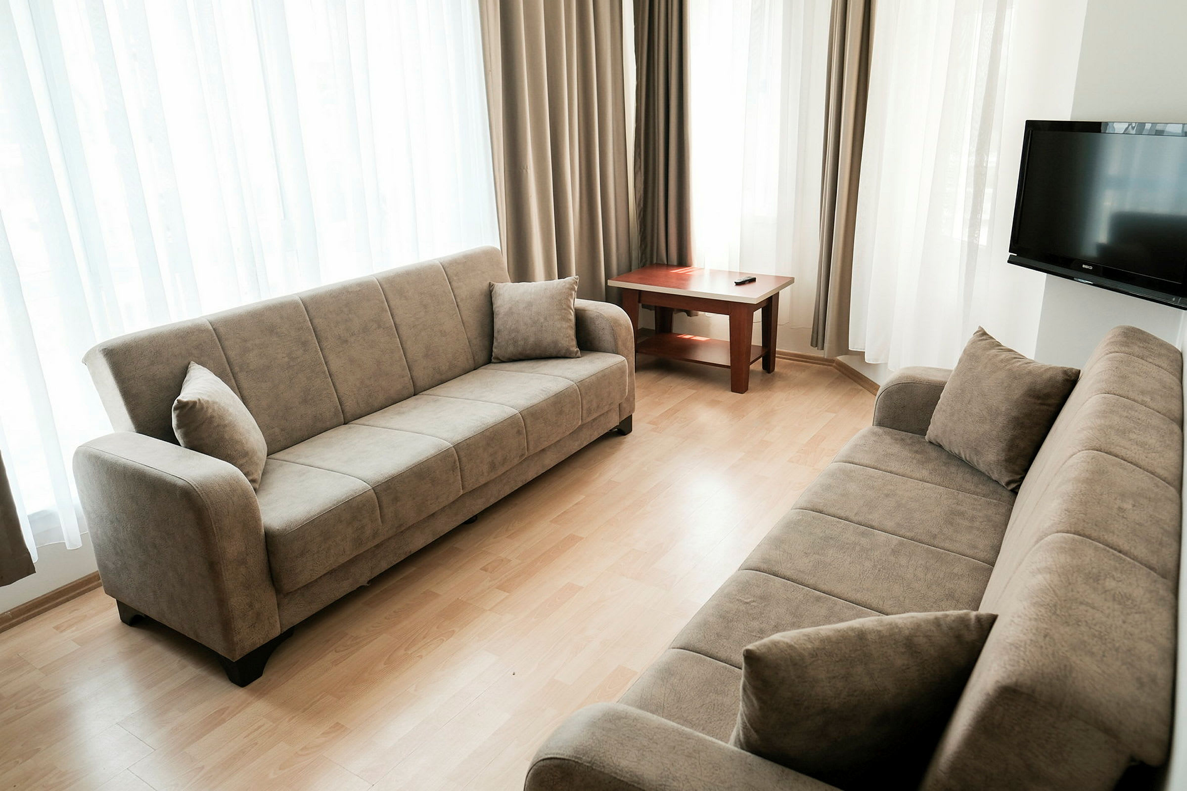 En stue med to beige sofaer, et træbord og et TV på væggen. Rummet har lyse gardiner ved vinduerne. (AI-genereret tekst)