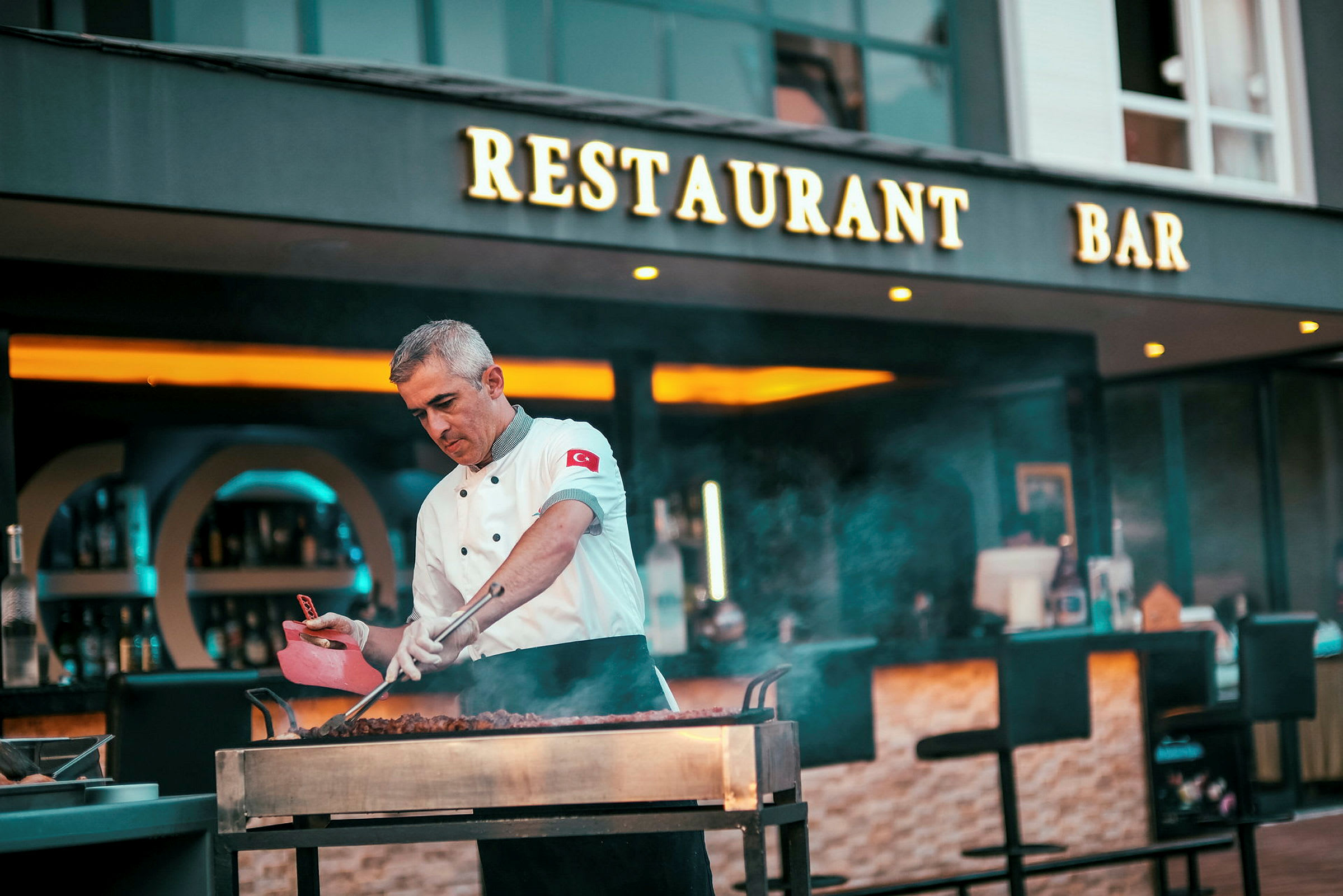 En kok tilbereder mad på en åben grill foran en restaurant og bar. (AI-genereret tekst)