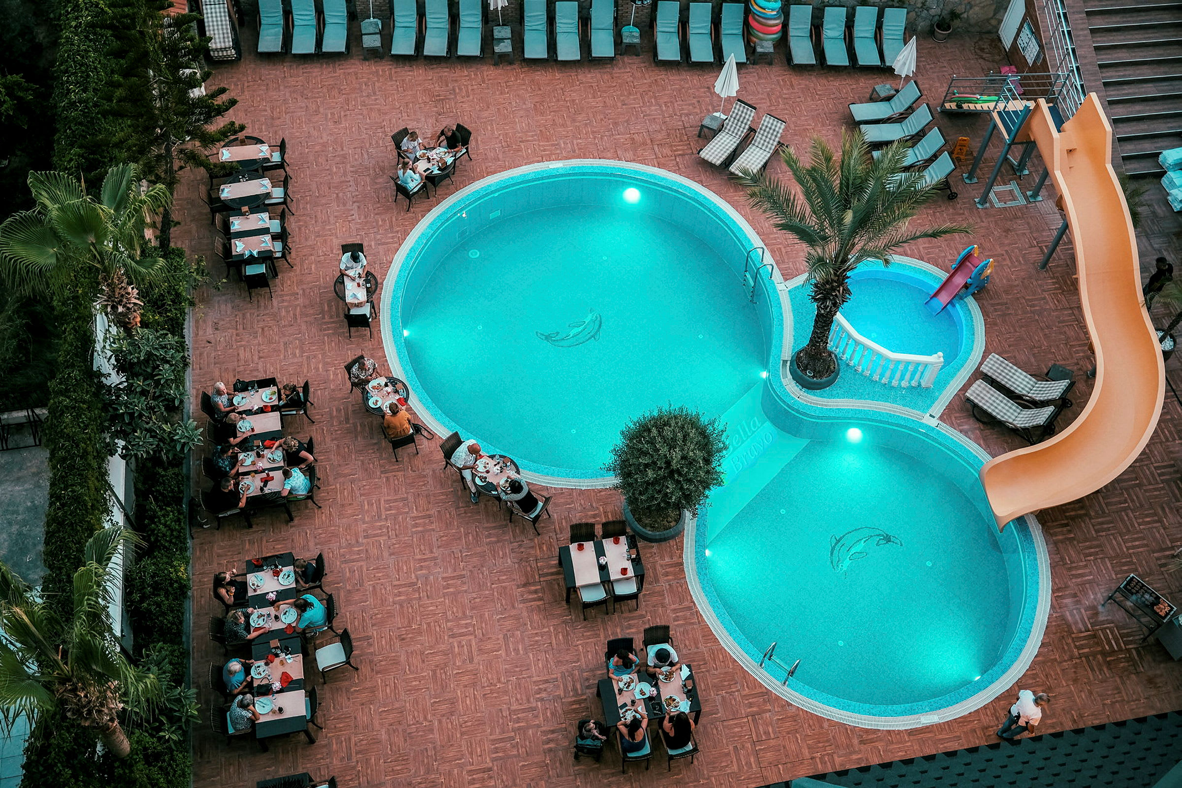 En udendørs pool på et hotelområde med liggestole og personer, der sidder ved borde og spiser. (AI-genereret tekst)