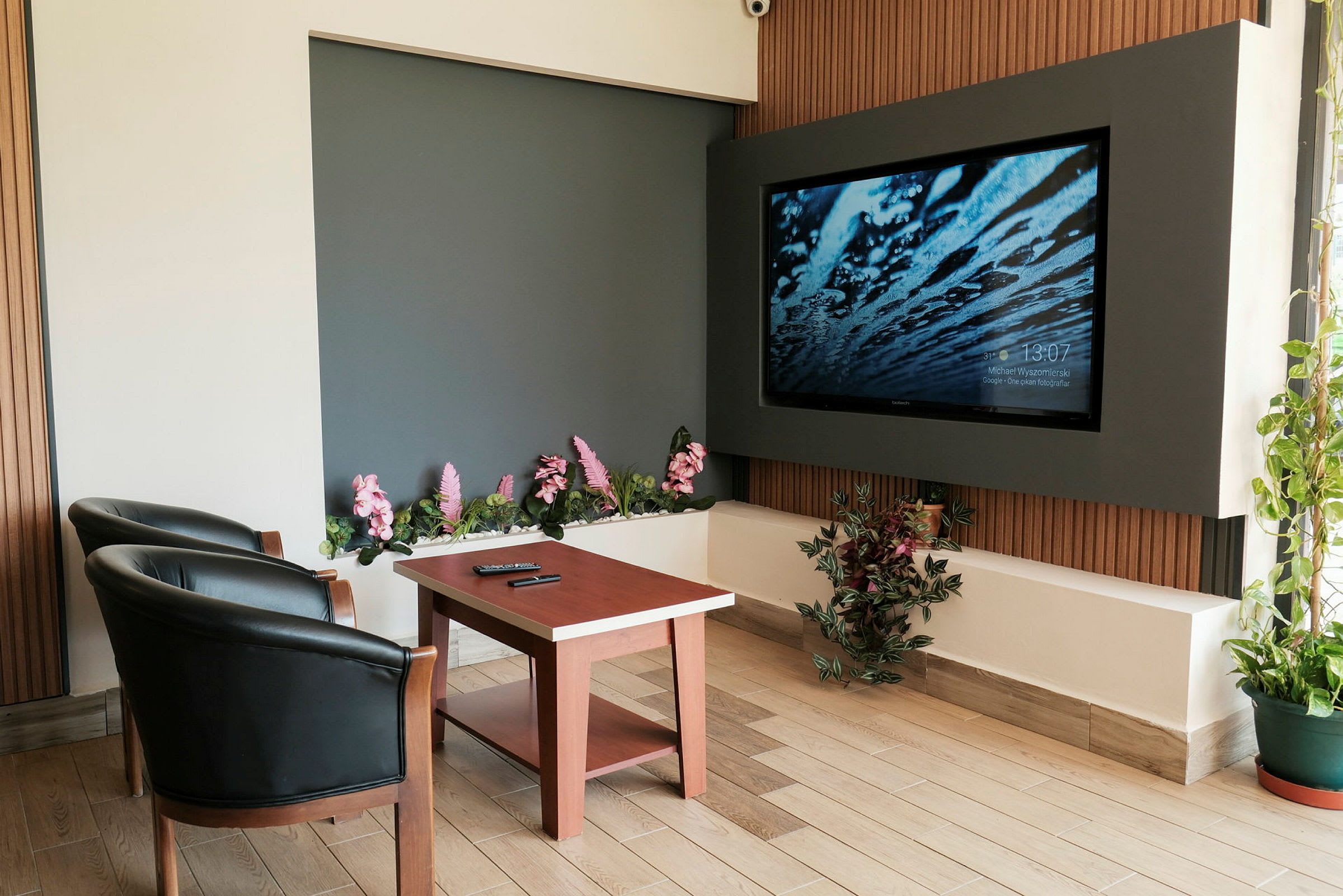 Et rum med to sorte lænestole, et træbord og et stort vægmonteret TV. Der er dekorative blomster og planter omkring. (AI-genereret tekst)