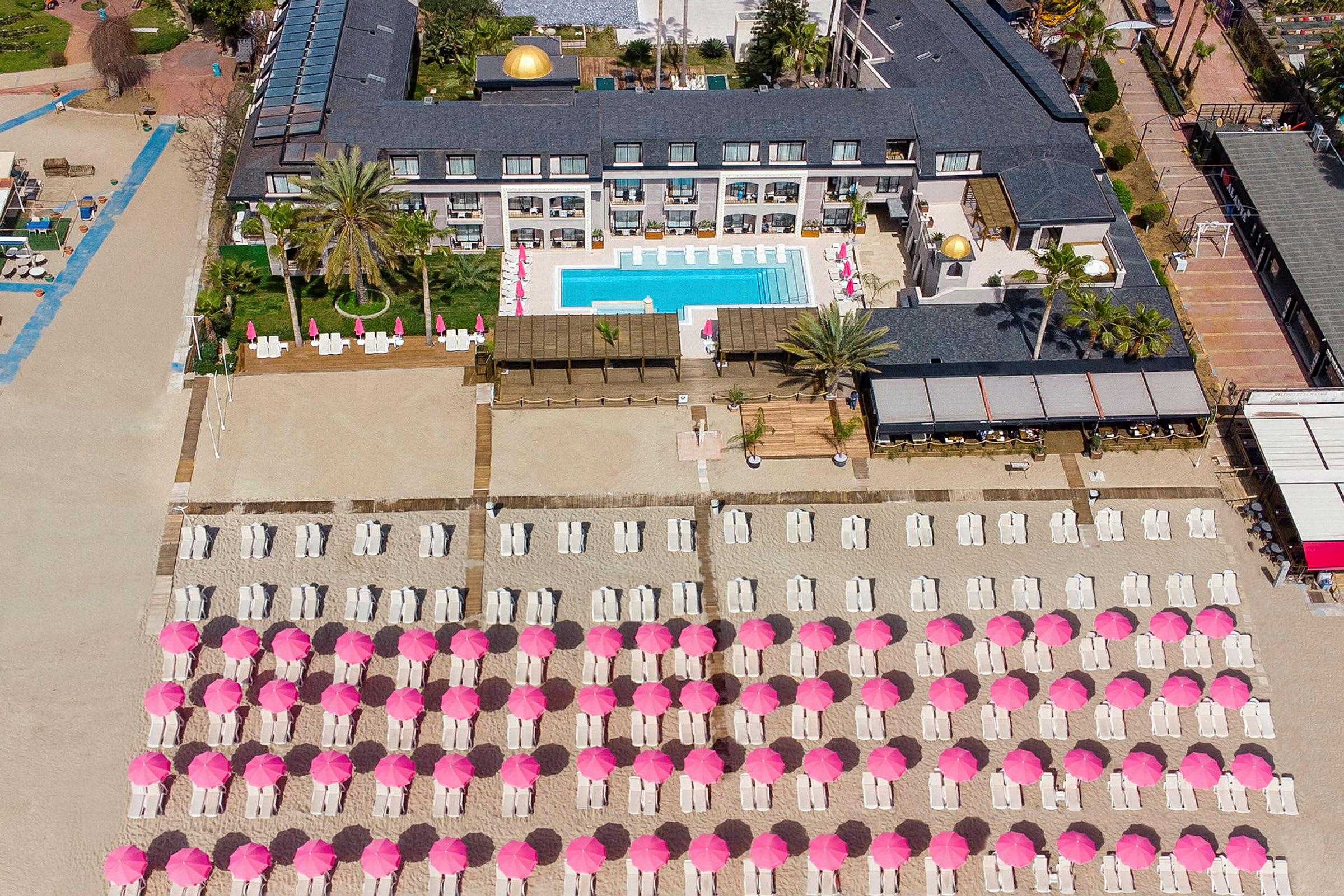 Fugleperspektiv over hotelområde med pool og liggestole under lyserøde parasoller på stranden. (AI-genereret tekst)