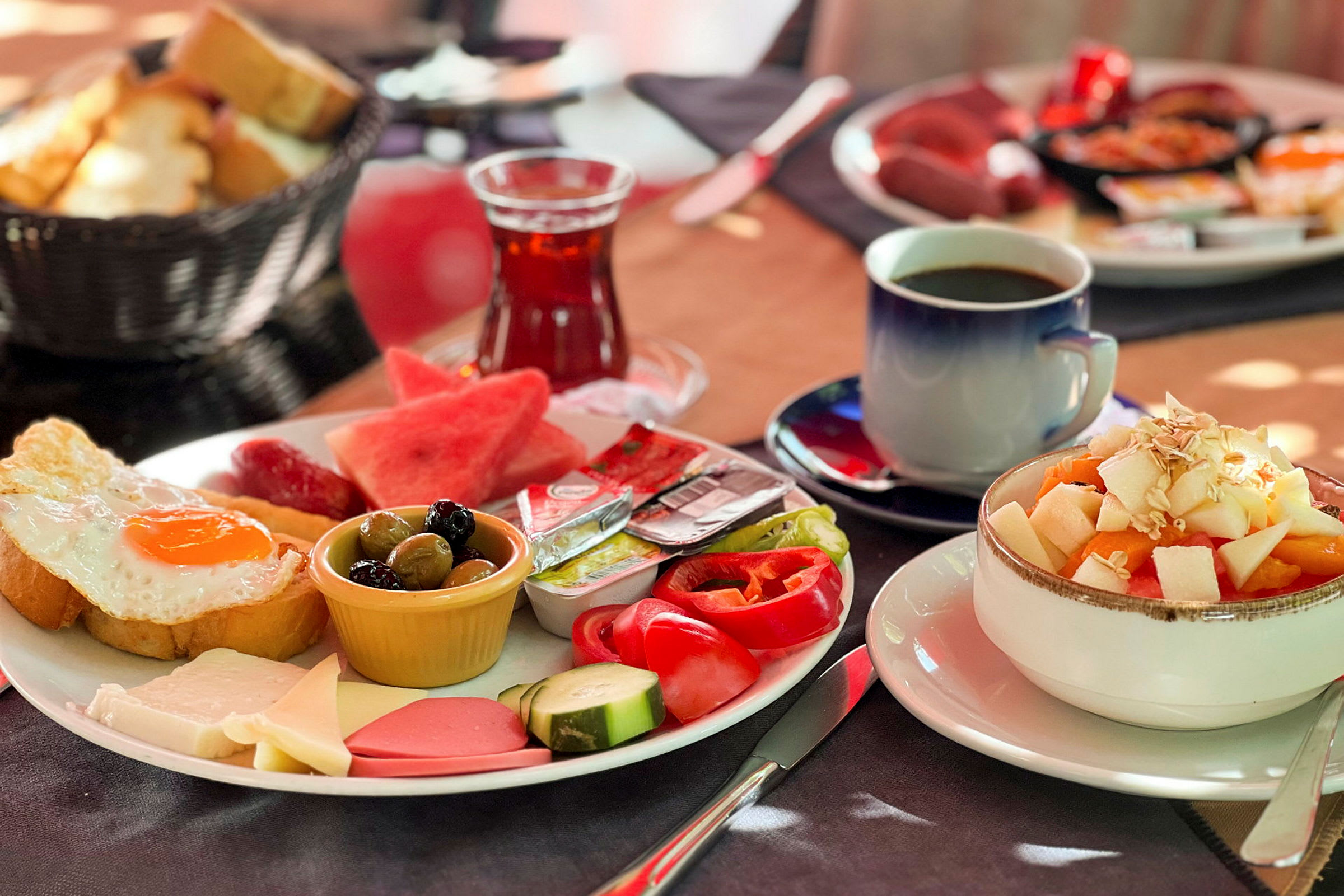 En tallerken med morgenmad, herunder stegt æg, grøntsager, ost og andre tilbehør, ved siden af en skål med frugtsalat og drikkevarer. (AI-genereret tekst)