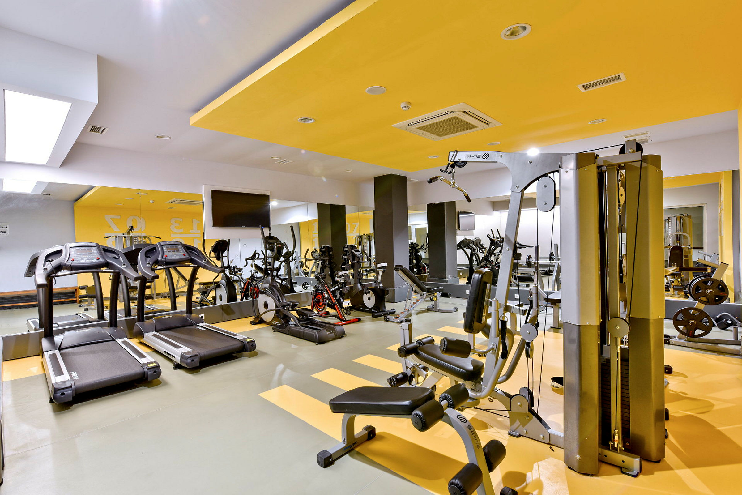 Et fitnesscenter med forskellige træningsmaskiner såsom løbebånd, cykler og styrketræningsudstyr. Væggene er malet i gul og hvid. (AI-genereret tekst)