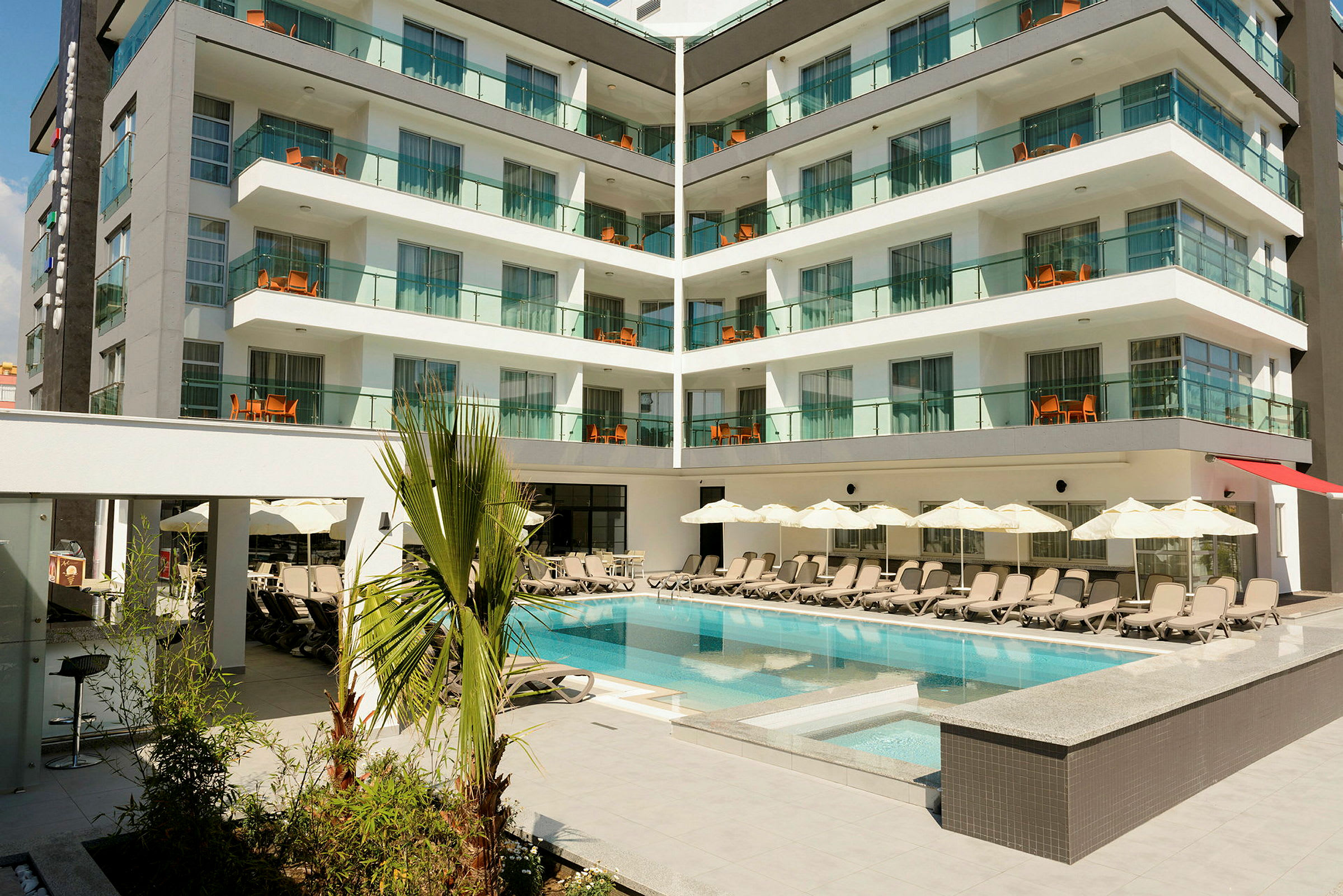 Hotelbygning med moderne altaner og en pool omgivet af liggestole og parasoller. (AI-genereret tekst)