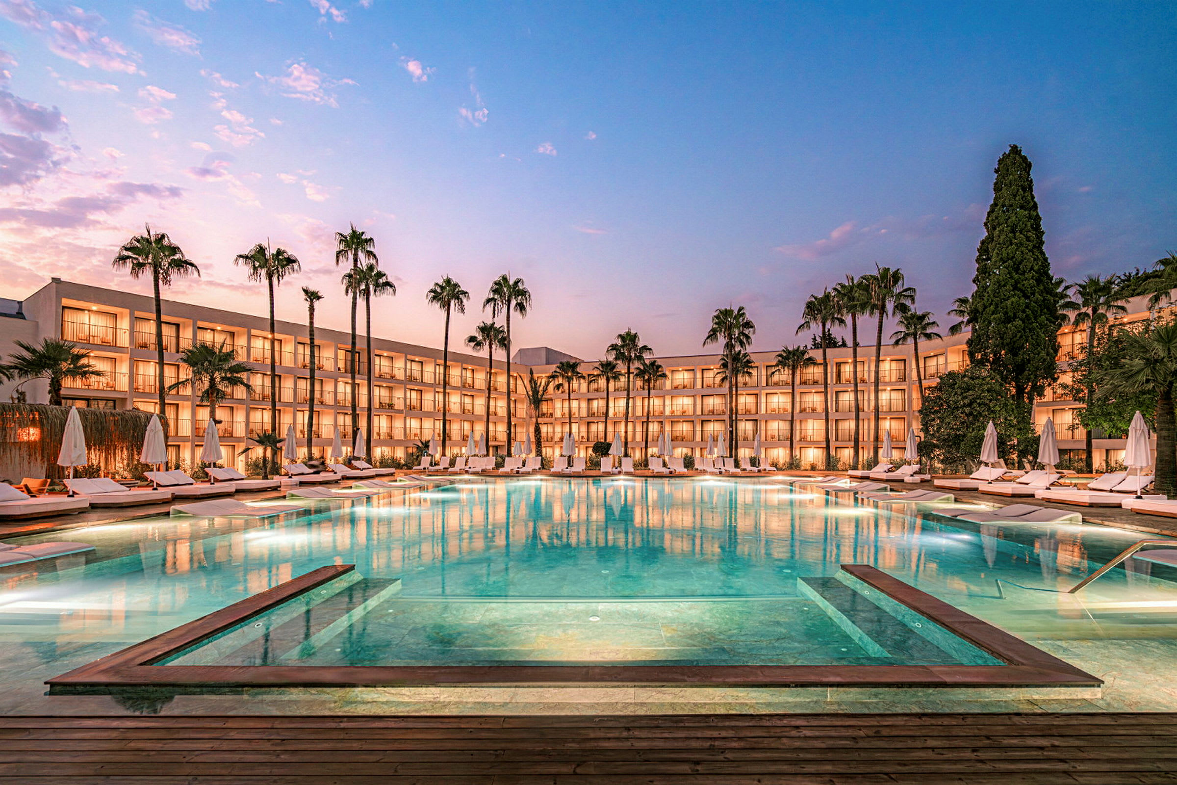 Aftenbillede af hotel med pool og omkringliggende liggestole. (AI-genereret tekst)