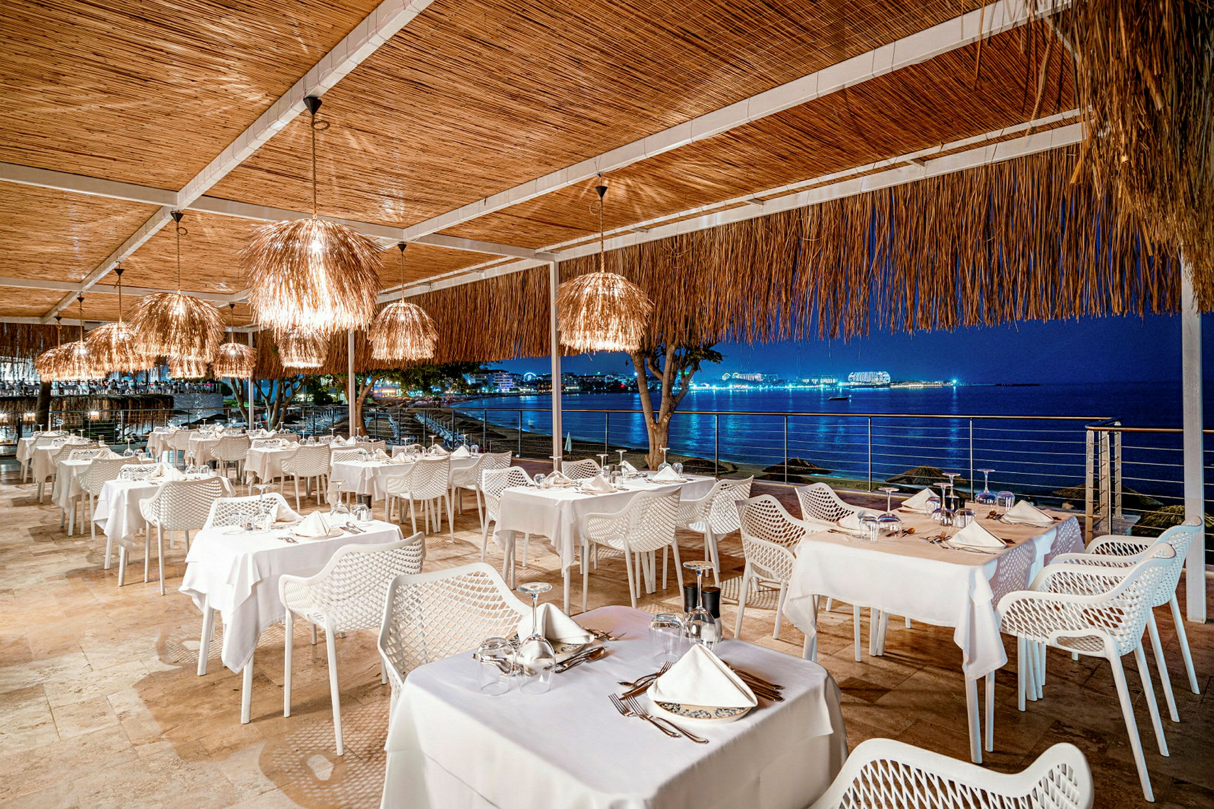 En udendørs restaurant med borde dækket med hvide duge og stole, og udsigt mod en oplyst havbugt om natten. (AI-genereret tekst)