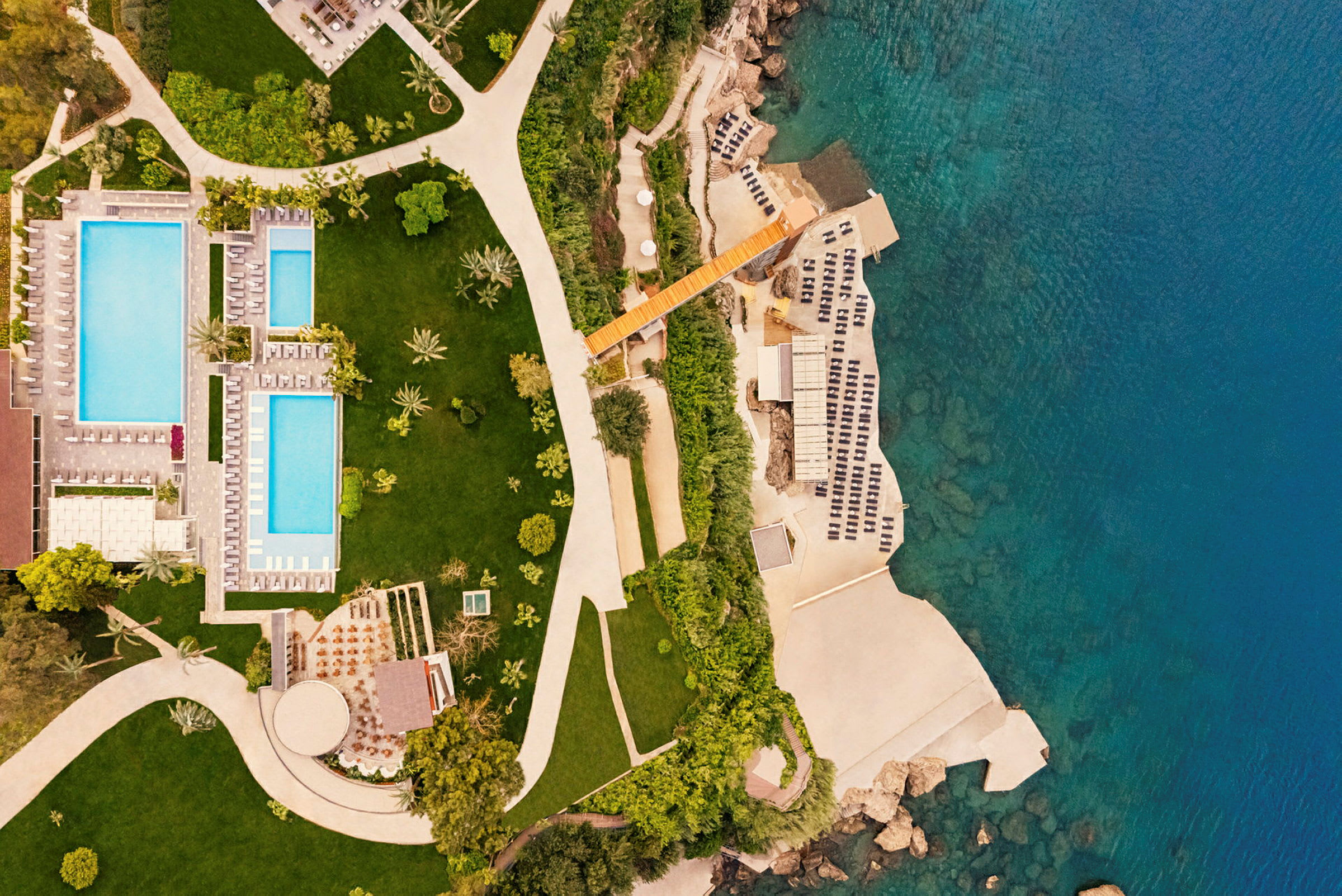Luftfoto af et hotelområde med pools, grønne områder og hav. (AI-genereret tekst)