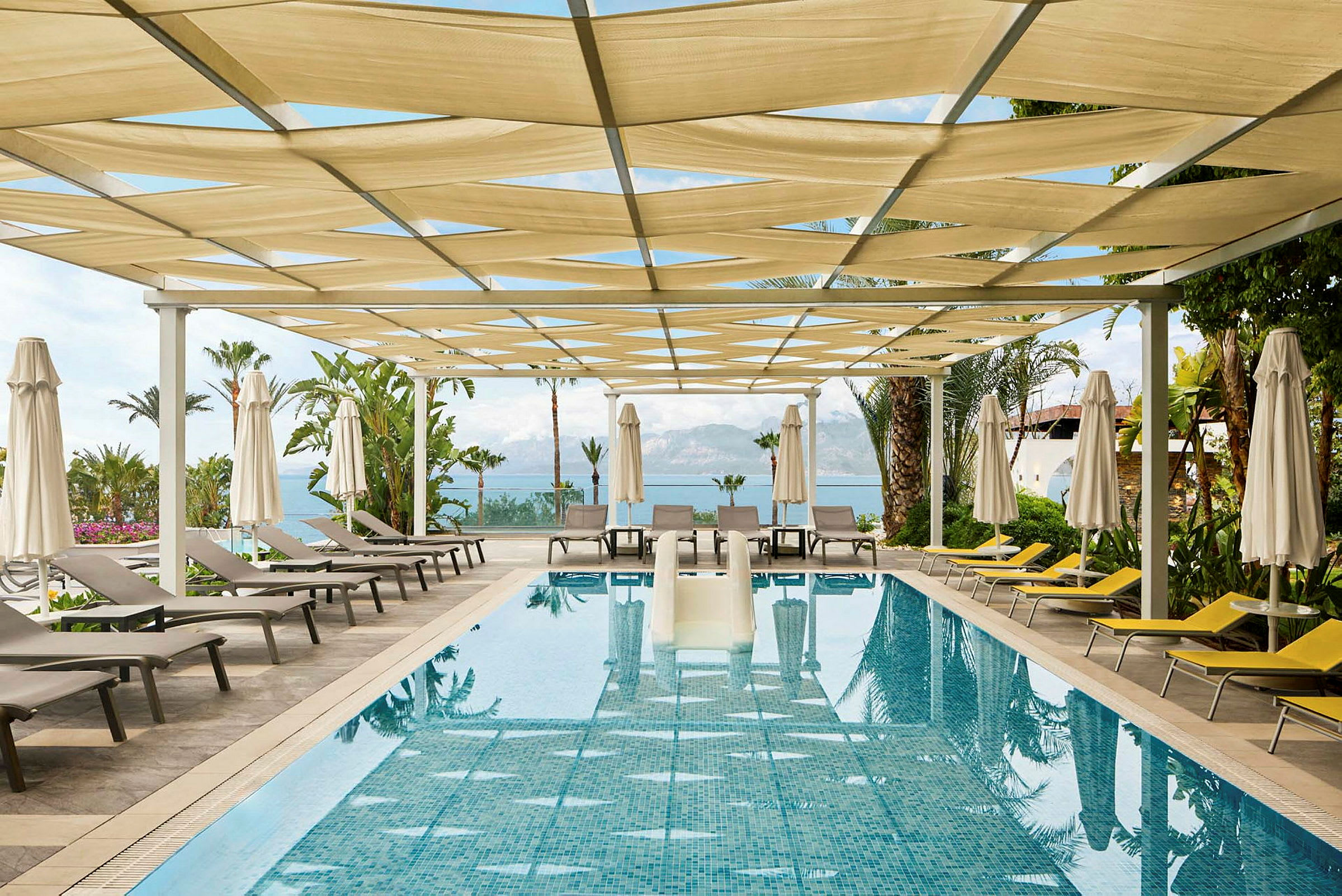 En swimmingpool med liggestole og parasoller under en pergola. (AI-genereret tekst)