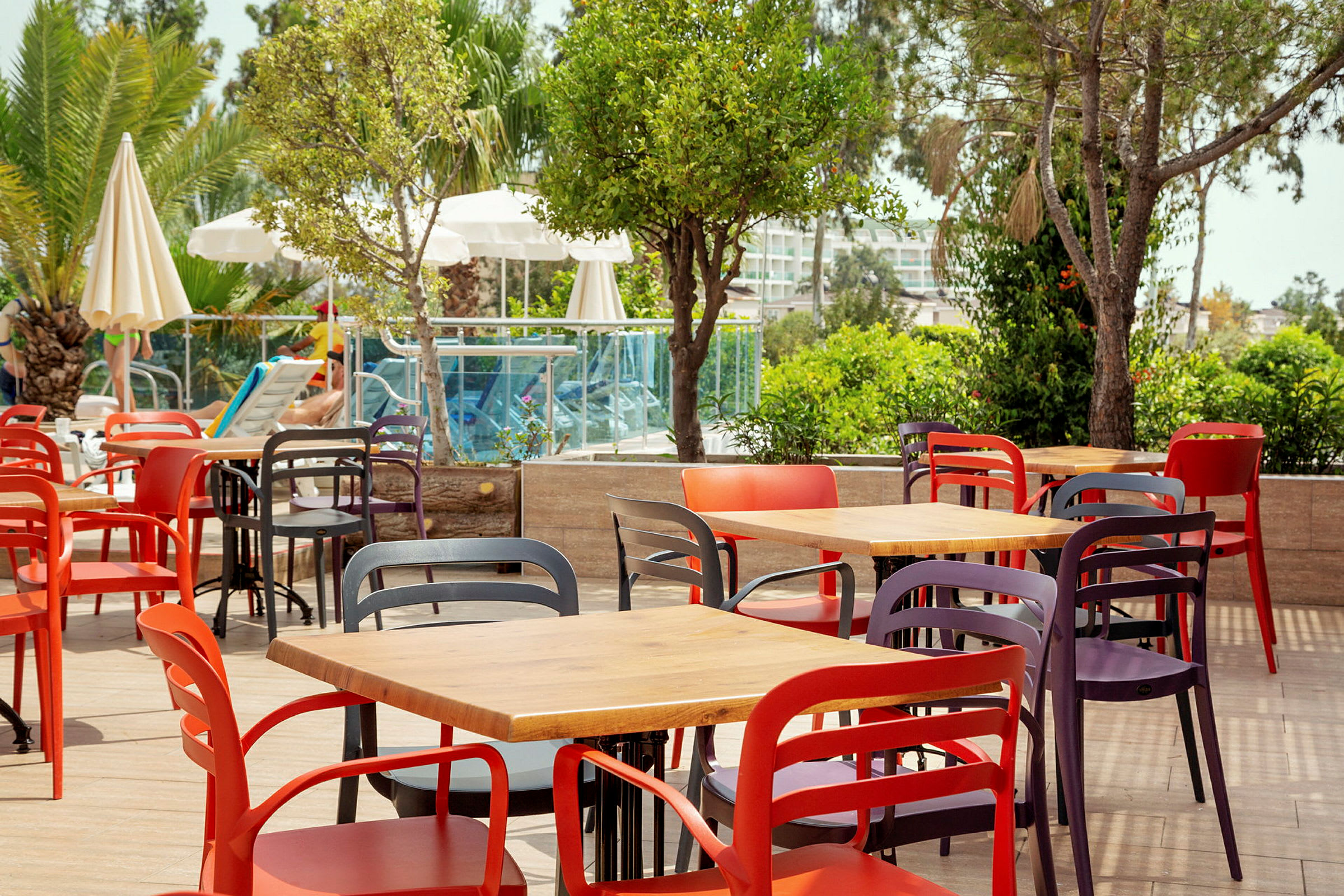 Farverige stole og borde arrangeret på en udendørs terrasse nær grønt og en pool. (AI-genereret tekst)