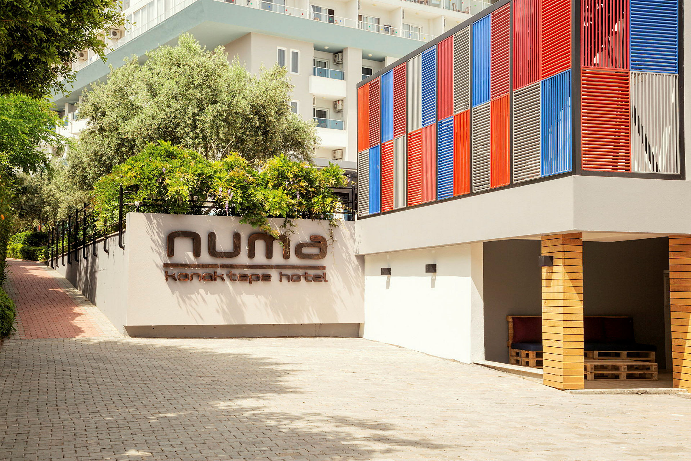 Hotellets facade med et skilt, der siger 'Numa Konaktepe Hotel', omgivet af planter og en farverig væg. (AI-genereret tekst)