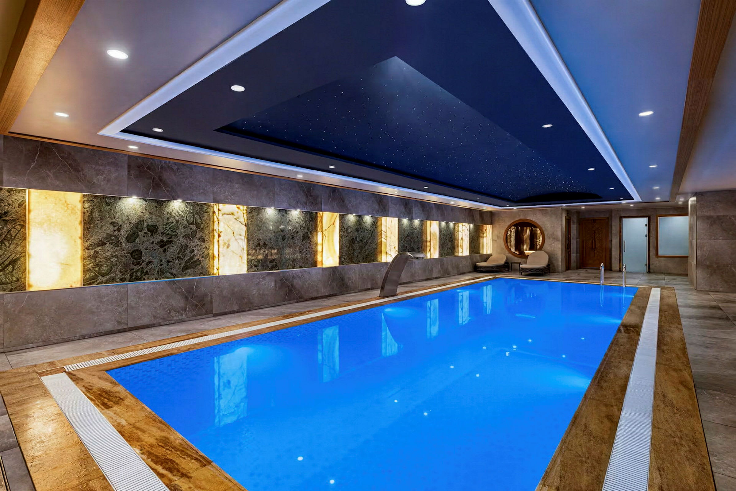 En indendørs pool med blåt vand i et elegant og moderne miljø. Væggene har dekorative paneler, og loftet har stjerneliknende lamper. (AI-genereret tekst)