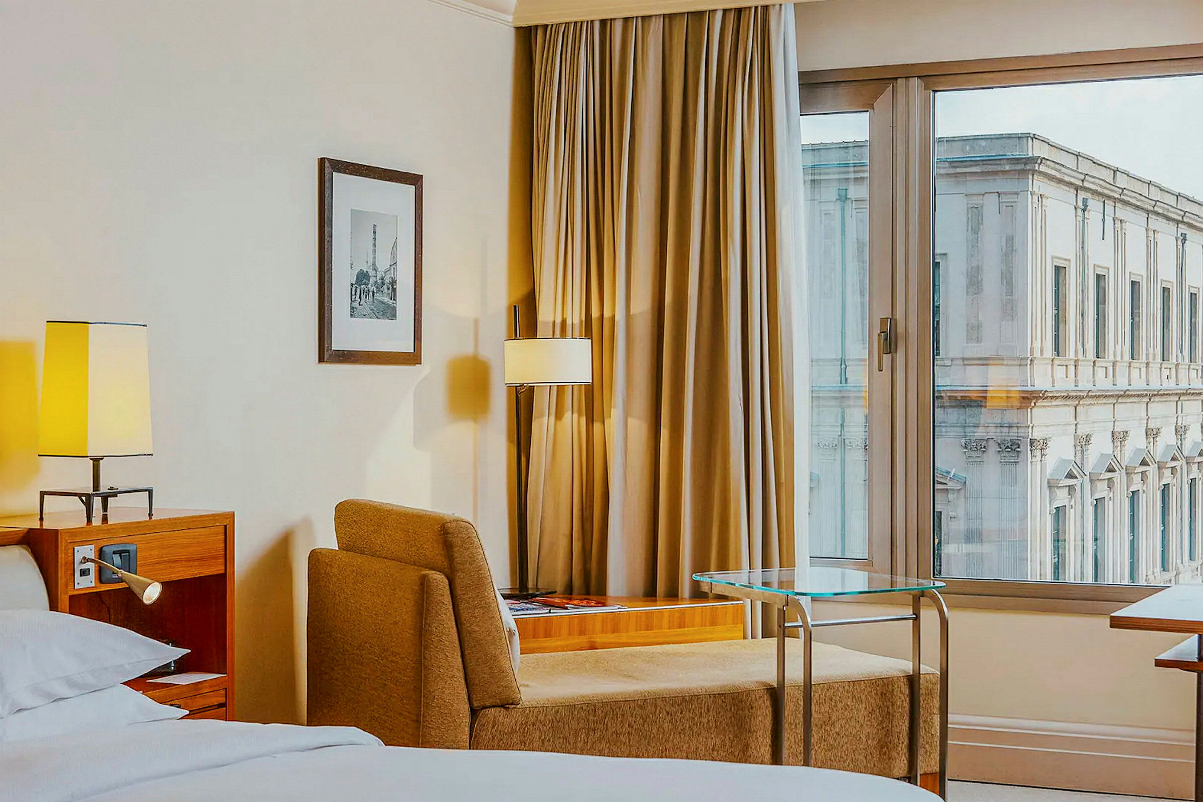 Hotelværelse med seng, lænestol, bord og vindue med udsigt til en bygning. (AI-genereret tekst)